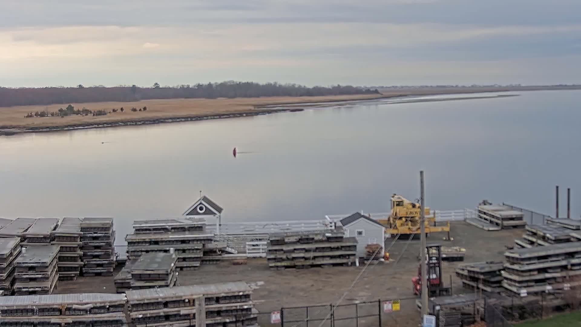 A calm, overcast day reveals a body of water with stacked floating docks and a crane near a small building, all situated next to a shoreline of muted brown grasses and distant trees.
