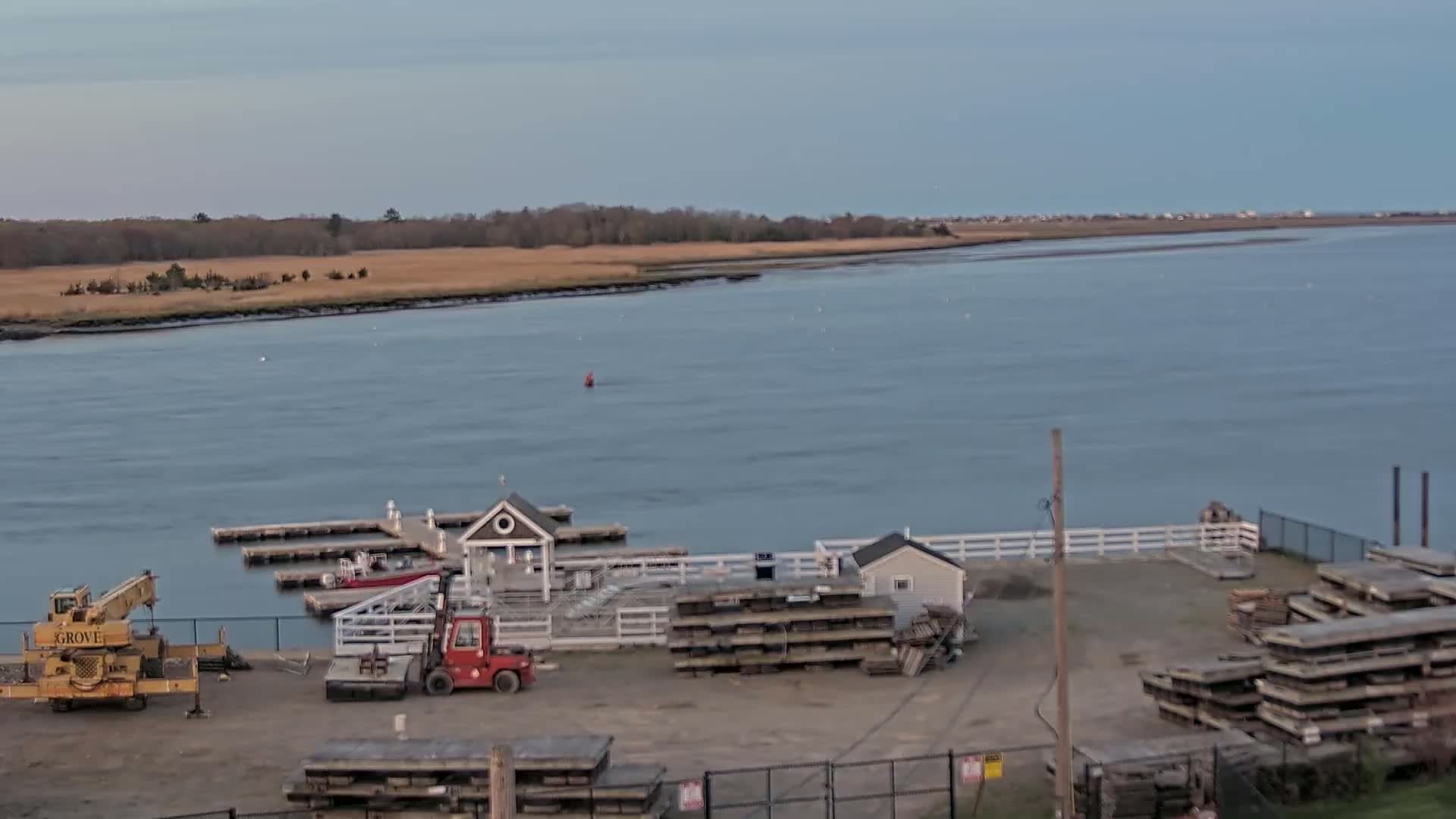 A calm body of water stretches toward a distant shoreline under a clear, pale sky, with a small dock, a crane, and stacks of lumber in the foreground.