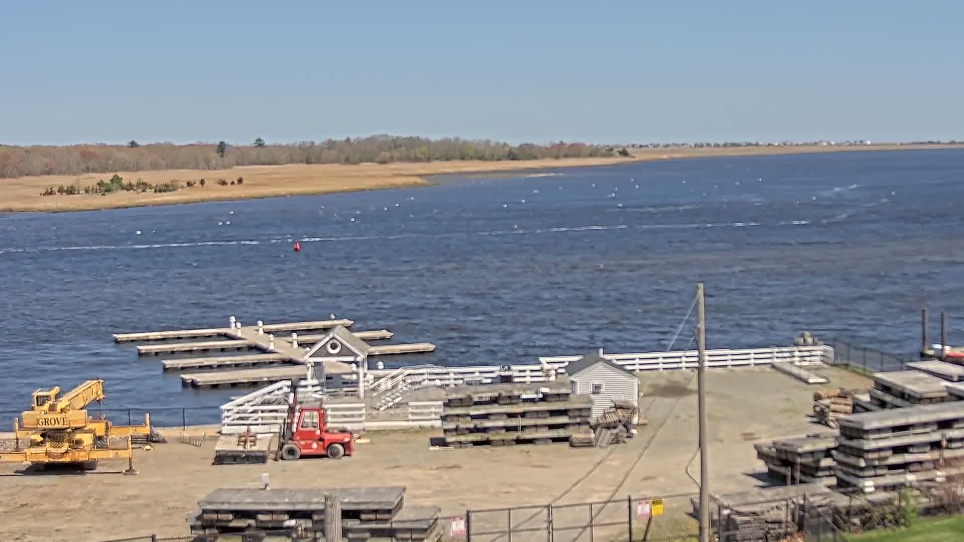 A body of water with a small marina, a yellow crane, a red forklift, and stacks of materials on a sunny day.