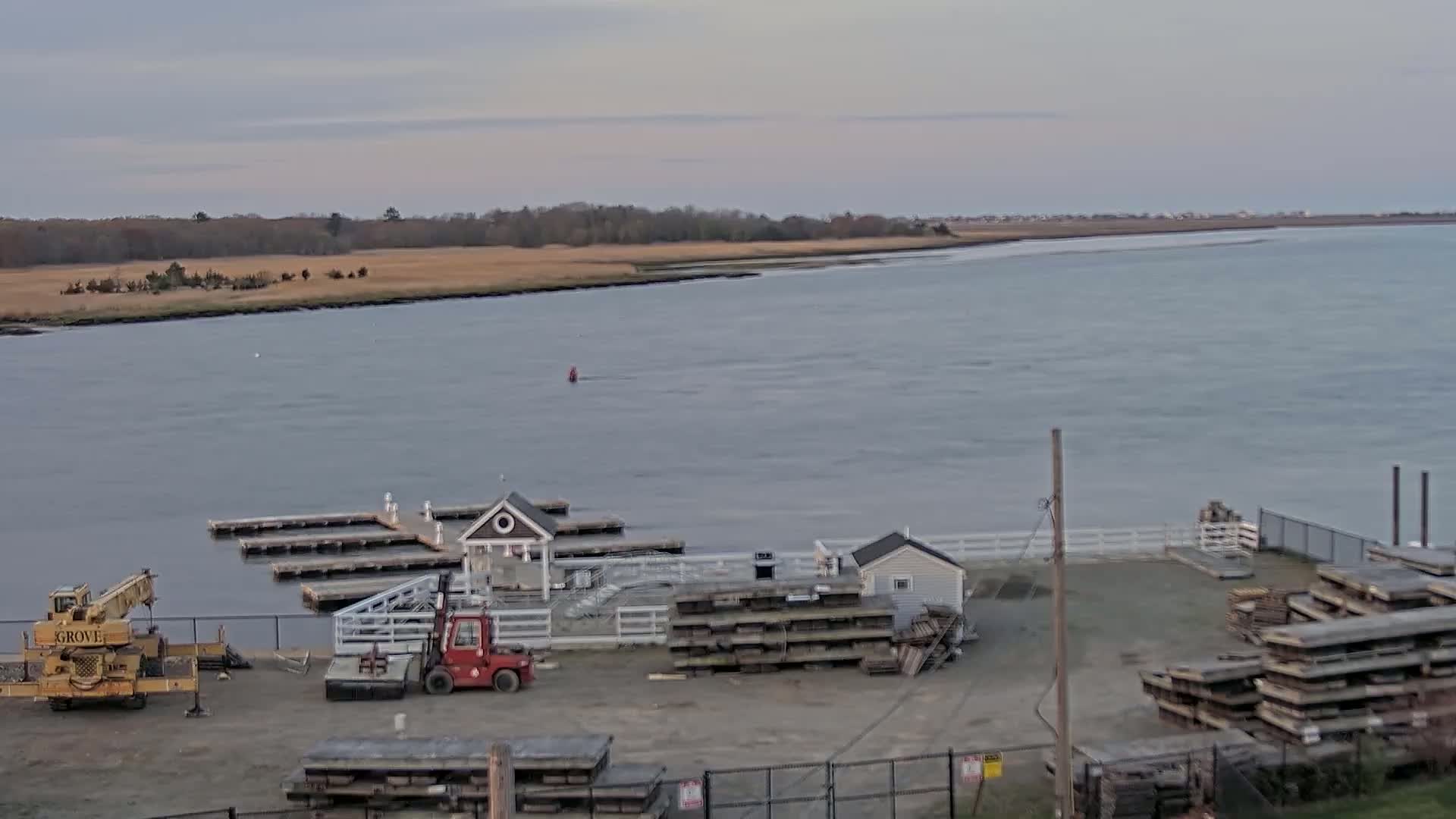 A calm, overcast day reveals a waterfront scene with a small marina, construction equipment, and stacks of lumber near a body of water bordered by marshland.