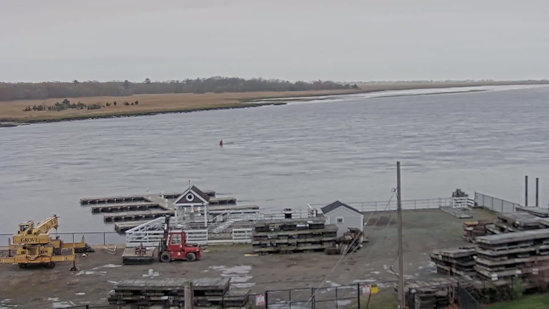 A marina with a small building and several stacks of lumber sits on the shore of a choppy, gray body of water under an overcast sky.