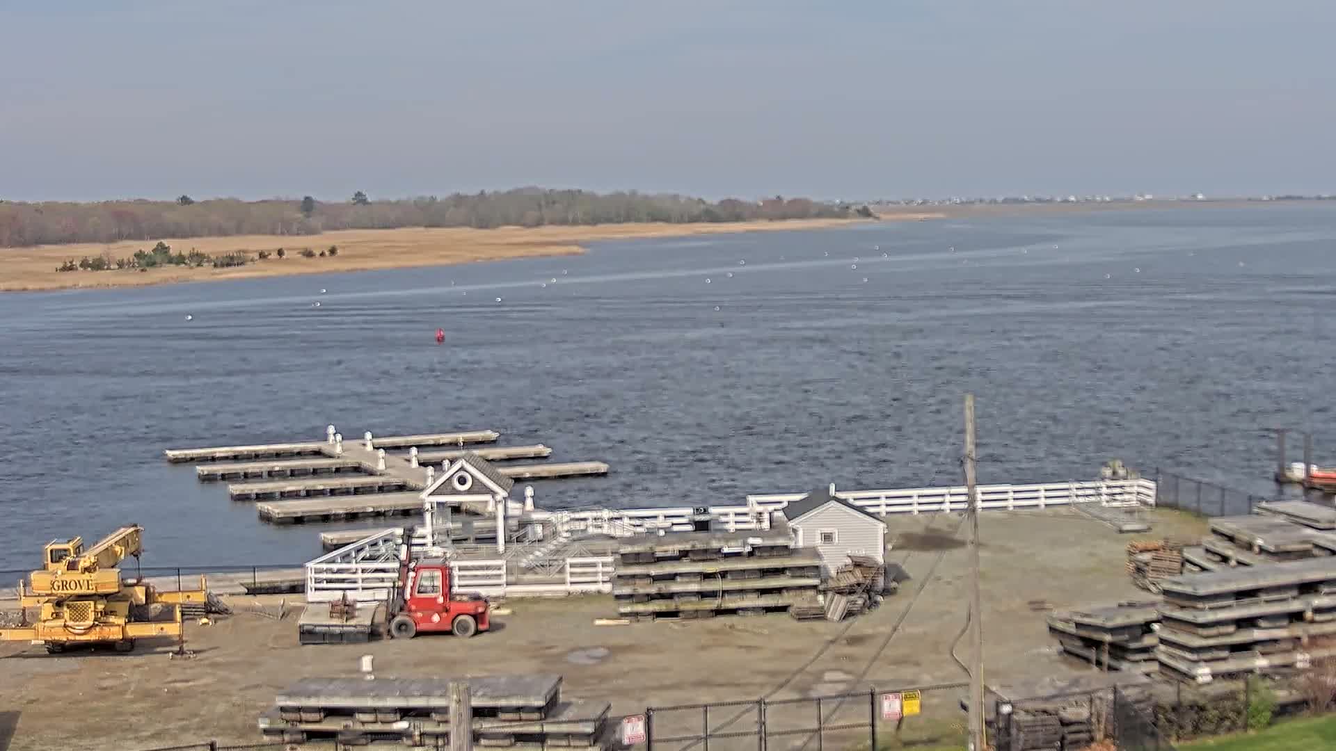 A mostly sunny day shows a marina with a small building, several docks, a yellow crane, and a red forklift, alongside a body of water with a shoreline visible in the distance.