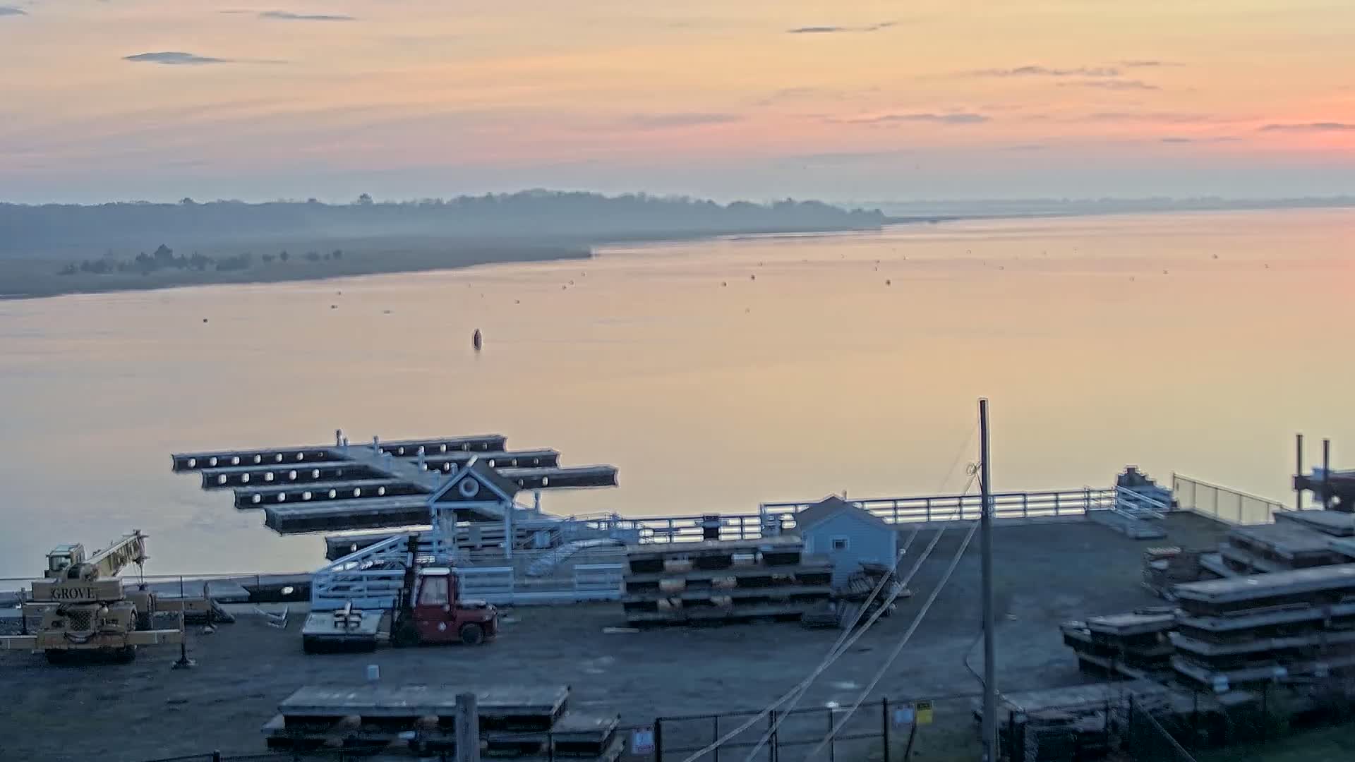 A calm, pink-hued sunrise over a wide body of water, showing a marina with several docks, a small building, and construction equipment.