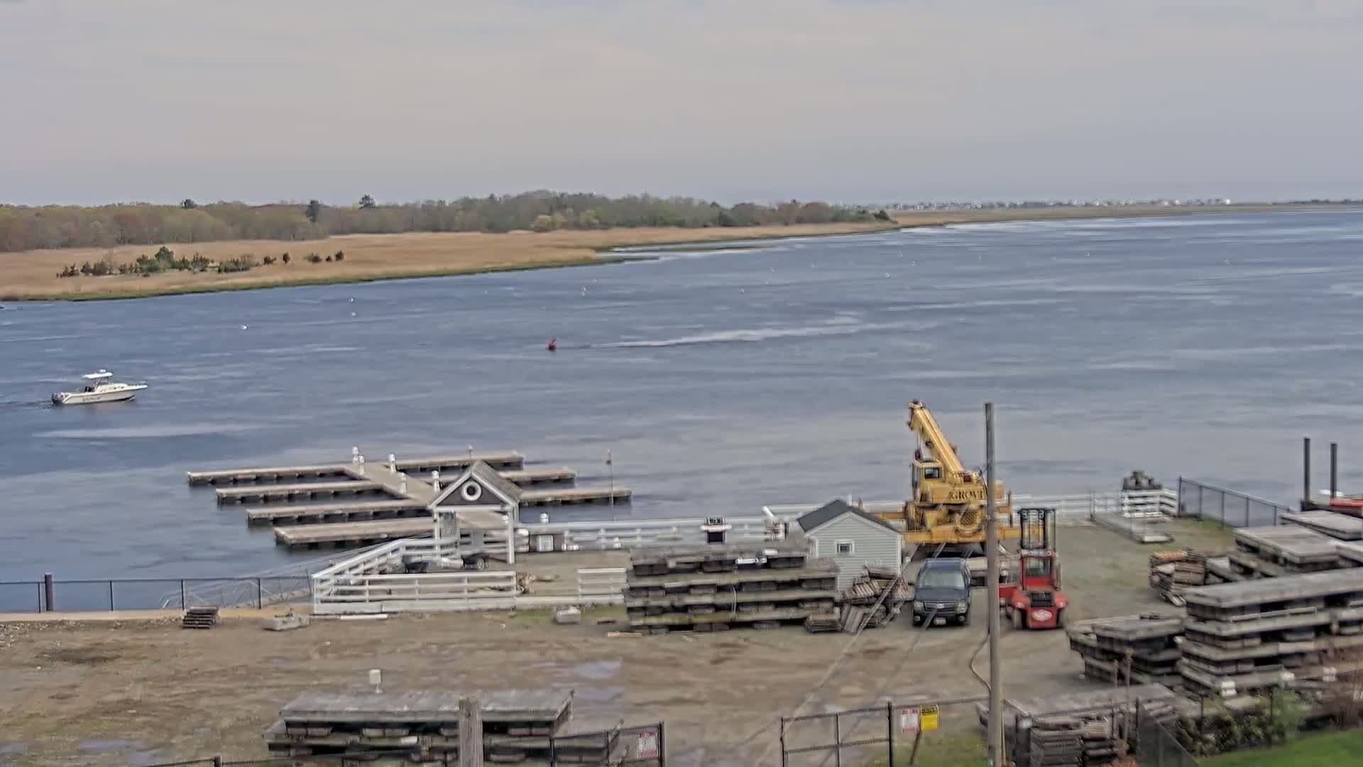 A body of water with a small boat and a marina is shown under an overcast sky, with a crane and stacks of materials on the shore.