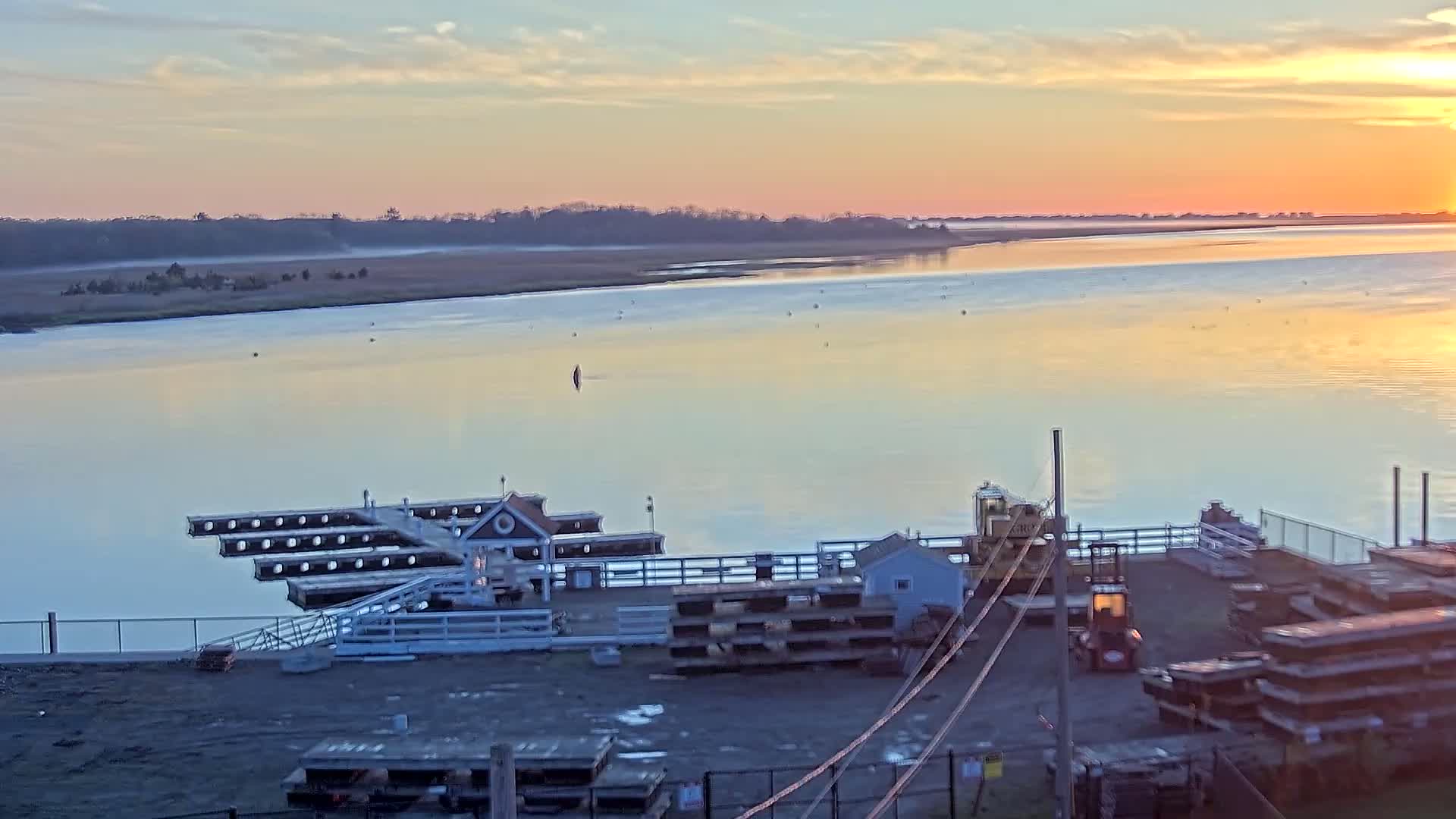 A calm, pale pink and orange sunrise illuminates a tranquil waterway with a small dock, a few buildings, and construction equipment.