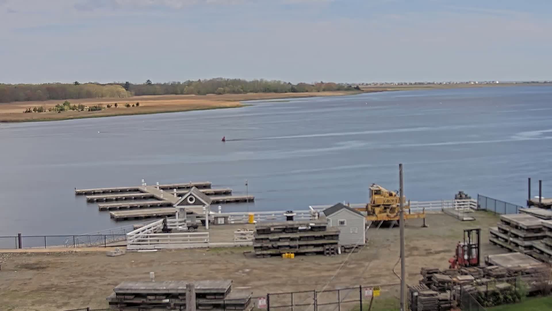 A calm body of water with a small marina, a crane, and stacks of materials on the shore under a partly cloudy sky.