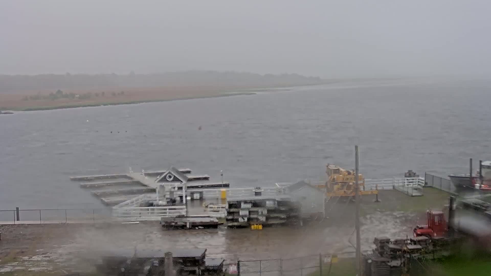 A small harbor with a building and docks is shown during a heavy rain, with choppy water visible in the background.