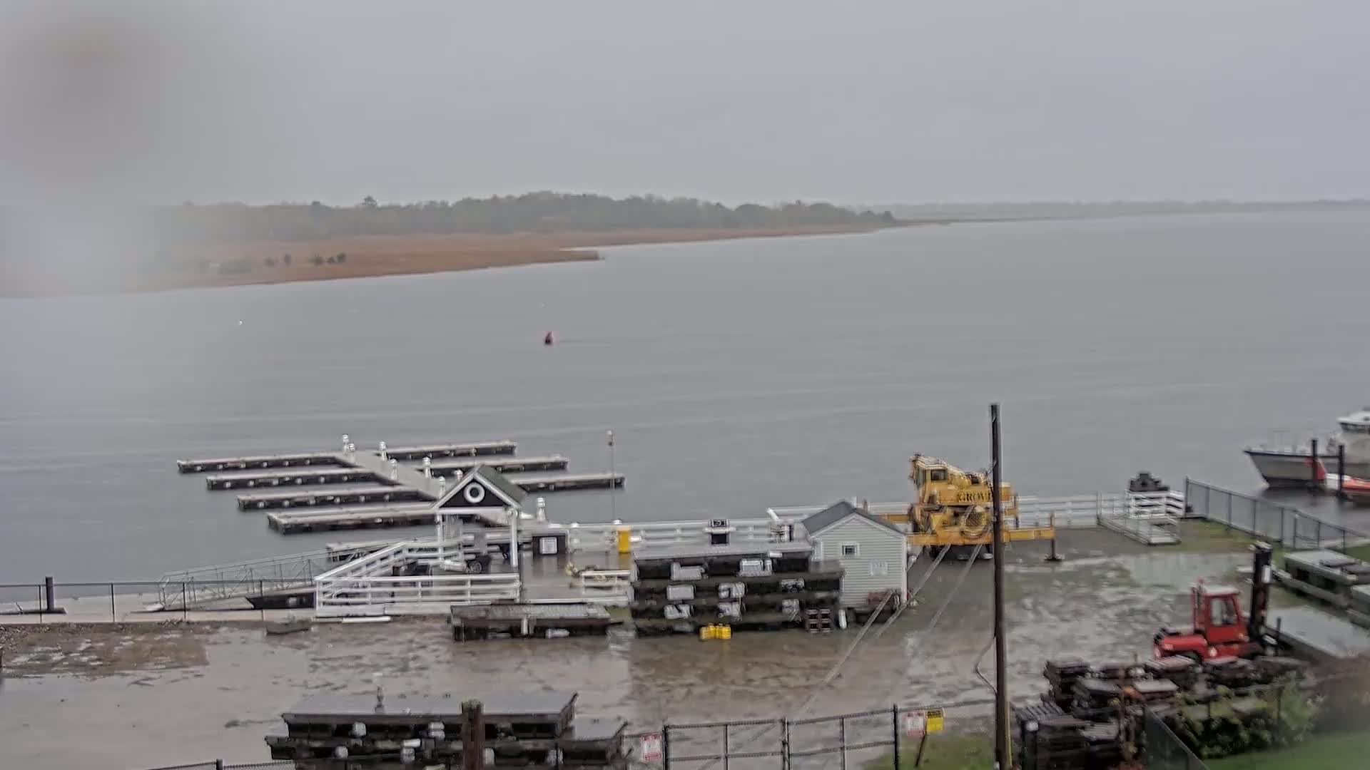 A marina with a small building and a crane is partially flooded under a gray, overcast sky, with a body of water extending to a distant shoreline.