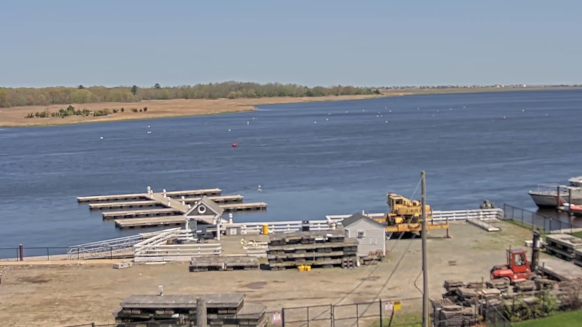 A marina with a small building and a yellow crane is situated on the shore of a calm, blue body of water under a clear, sunny sky, with a treeline visible in the distance.