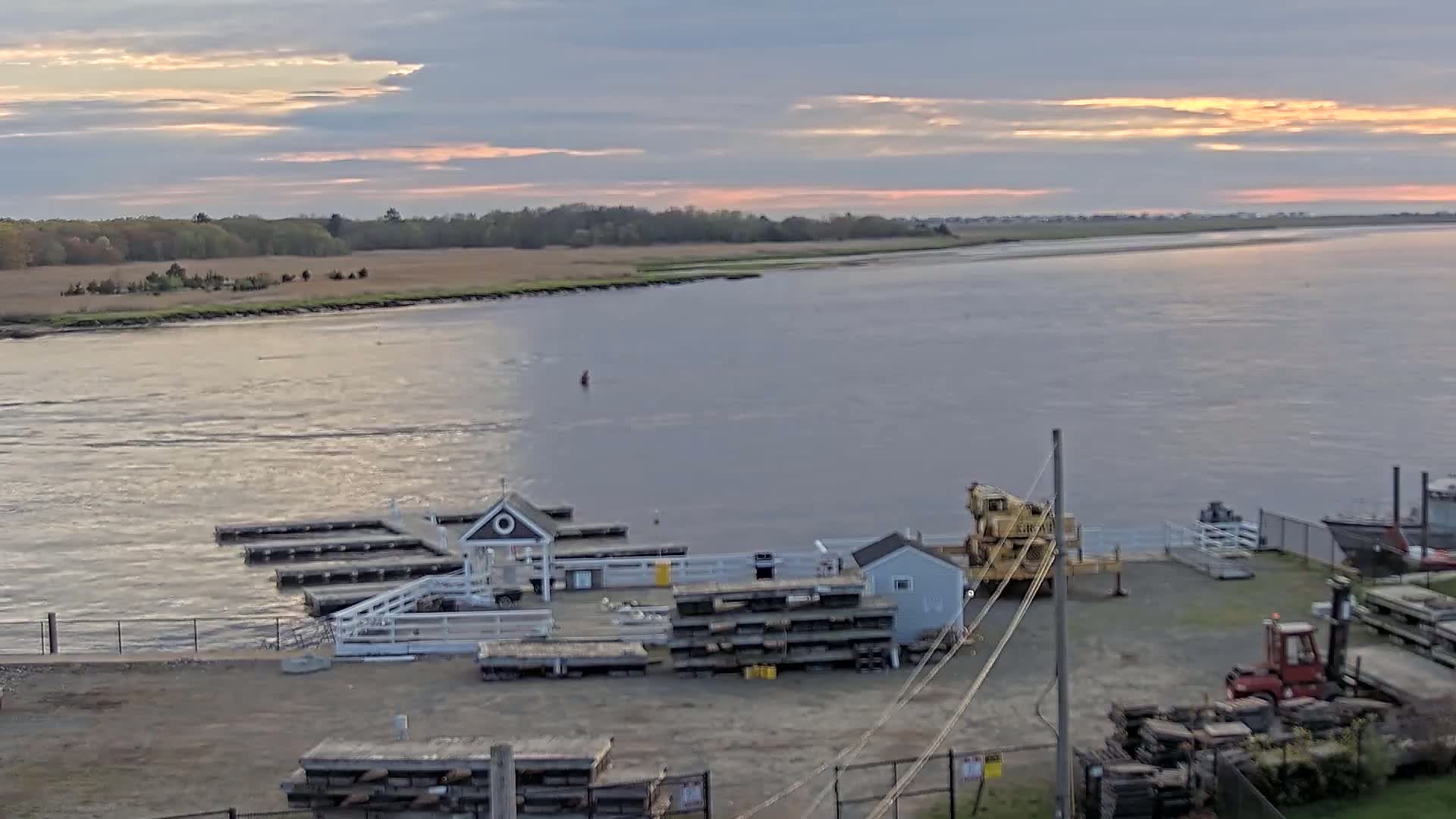 A calm river at sunset, with a small dock, several buildings, a crane, and a forklift situated near the water's edge, and a grassy area visible across the river.