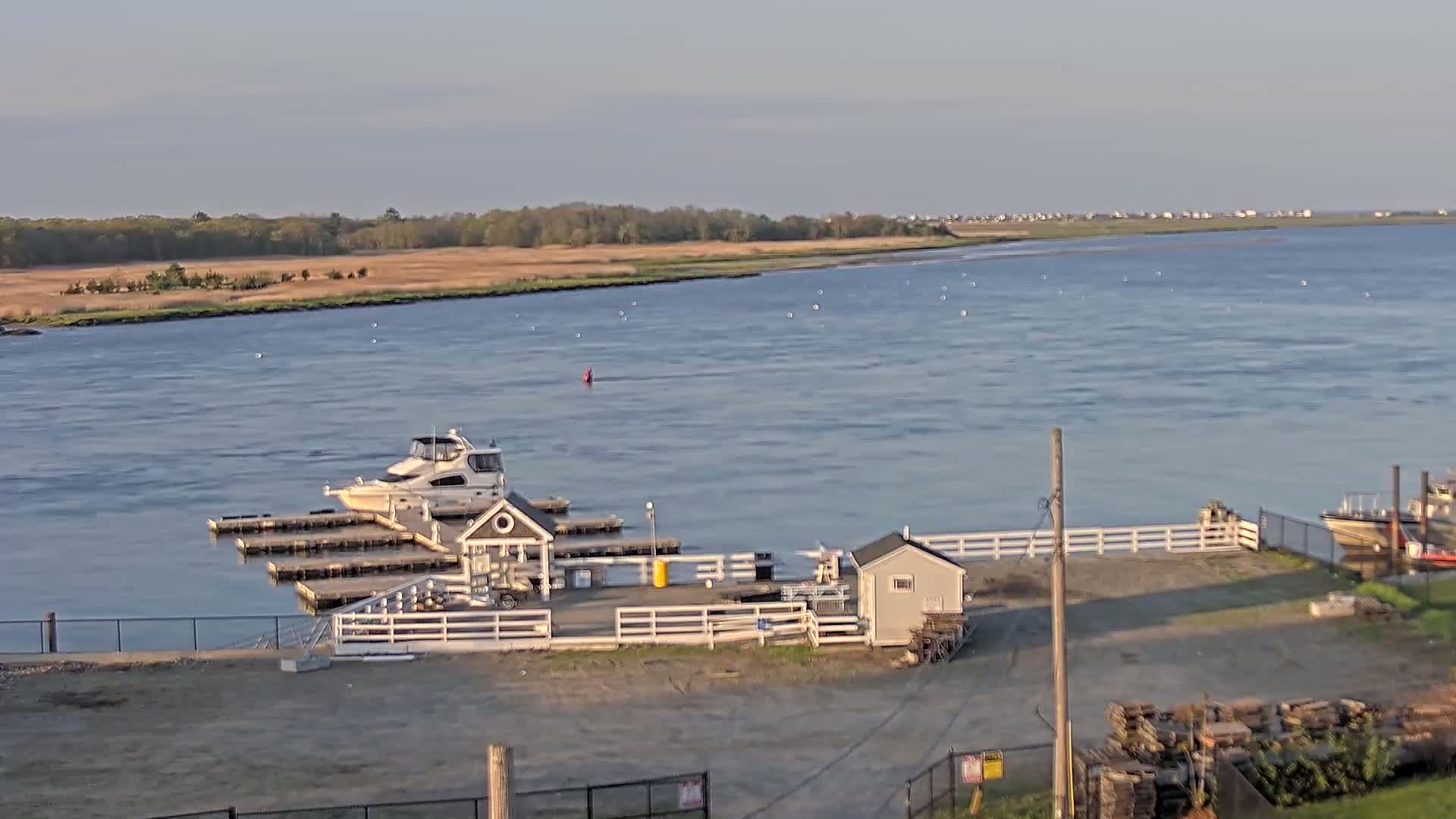 A calm river with a docked boat and small buildings near a shoreline with grassy marsh land under a clear sky.