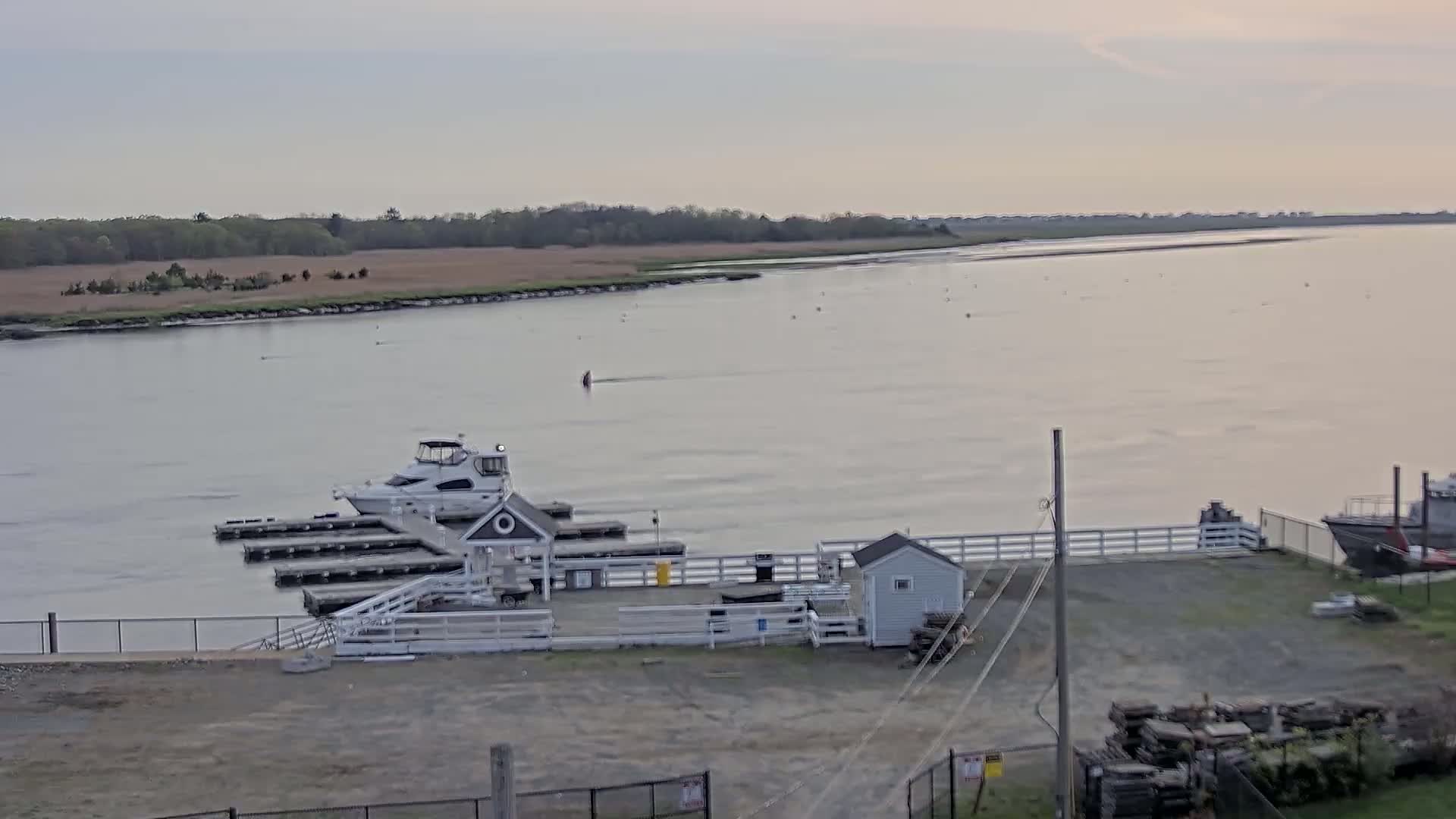 A calm, overcast day reveals a small marina with boats docked, a small building, and a larger body of water extending to a distant shoreline.