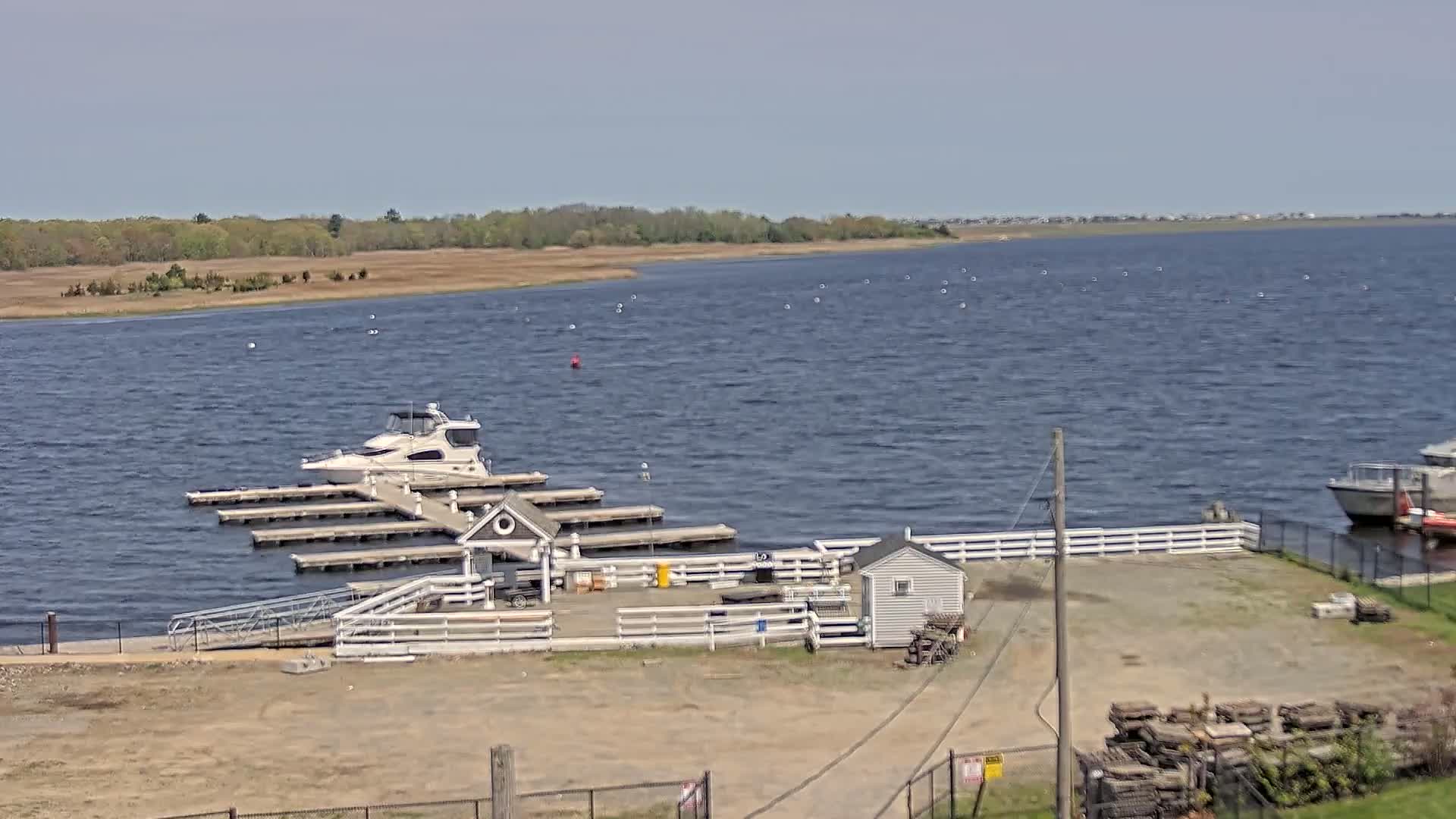 A small marina with a boat docked at multiple piers, a small building, and a larger boat in the distance on a partly sunny day with a slightly choppy bay.