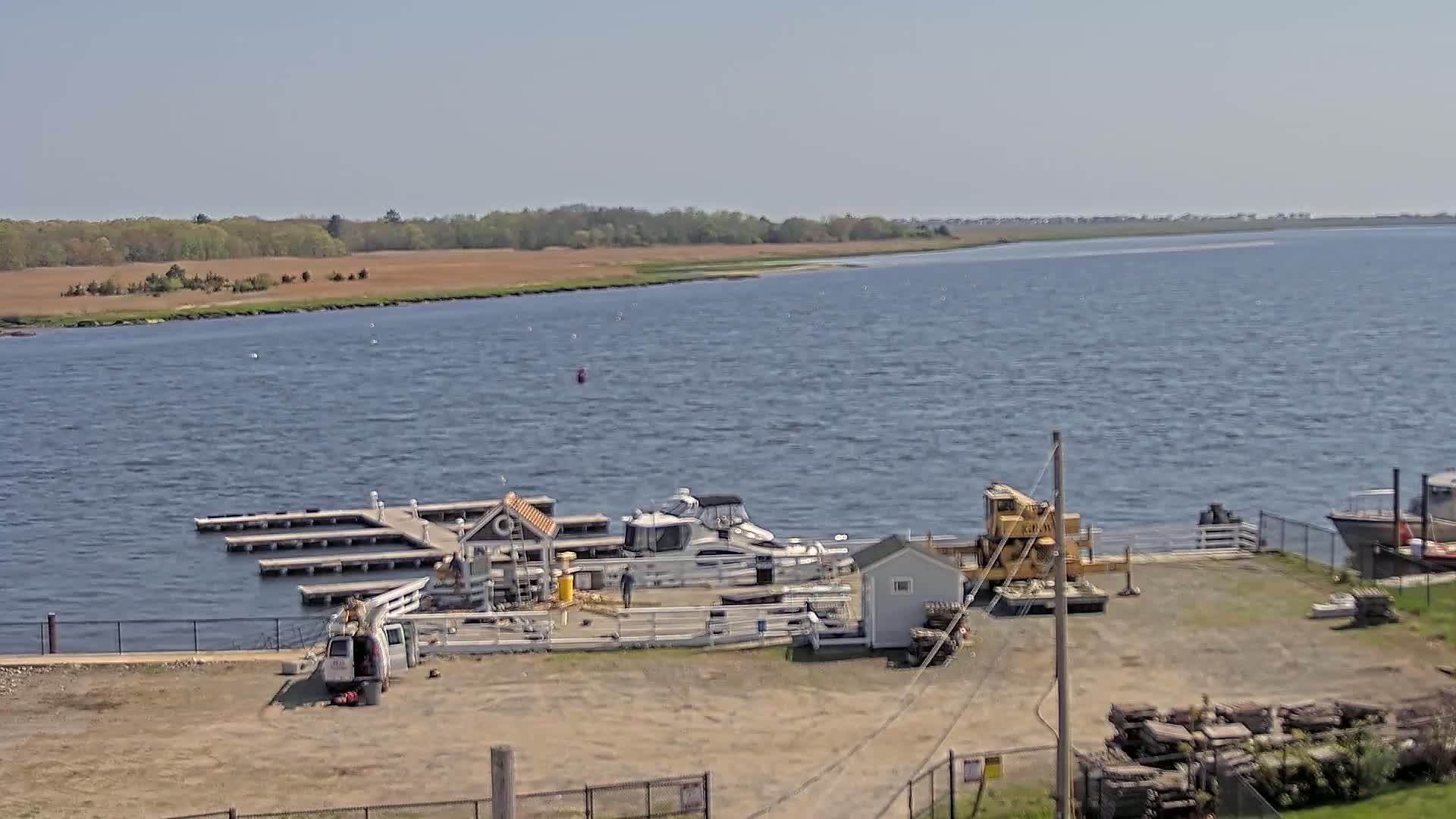 A small marina with boats docked, a yellow crane, and a few small buildings sits on the shore of a calm body of water under a clear, sunny sky.
