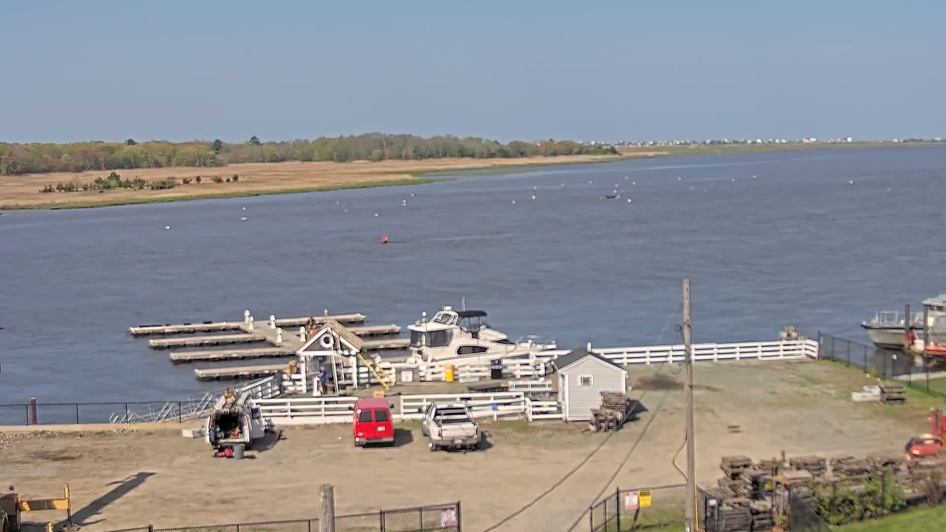 A small marina with a docked boat, several vehicles, and a small building sits on the edge of a calm body of water under a clear blue sky.