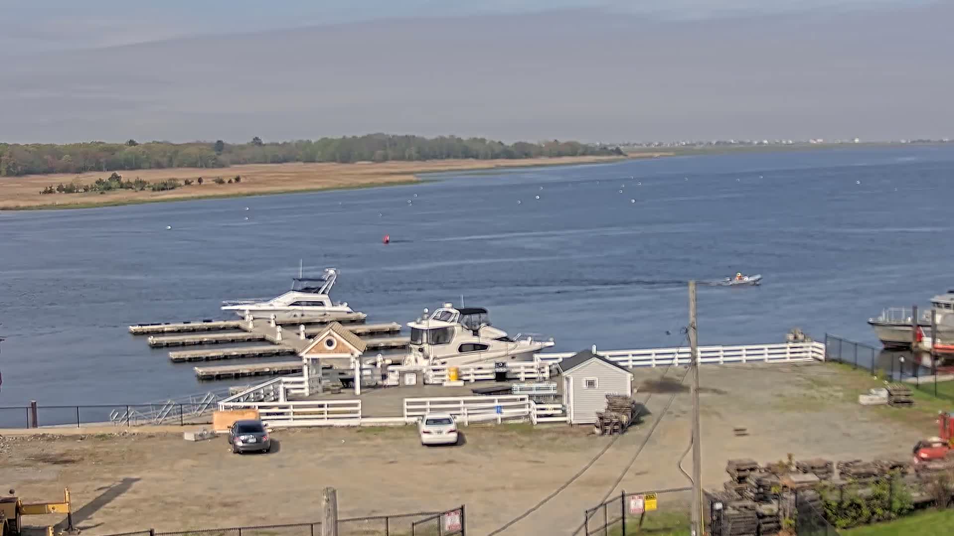 Several boats are docked at a marina on a calm, mostly sunny day, with a small motorboat on the water in the distance and a larger vessel further out.