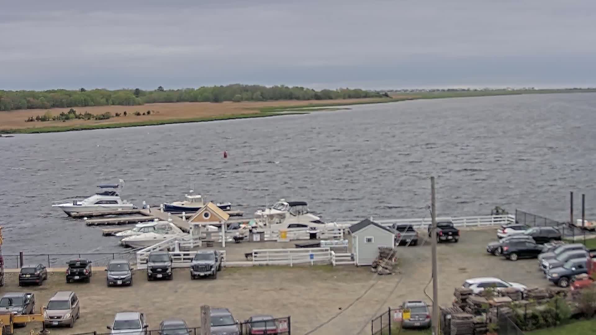 A marina with several boats docked and a parking lot full of cars is shown on a breezy, overcast day.