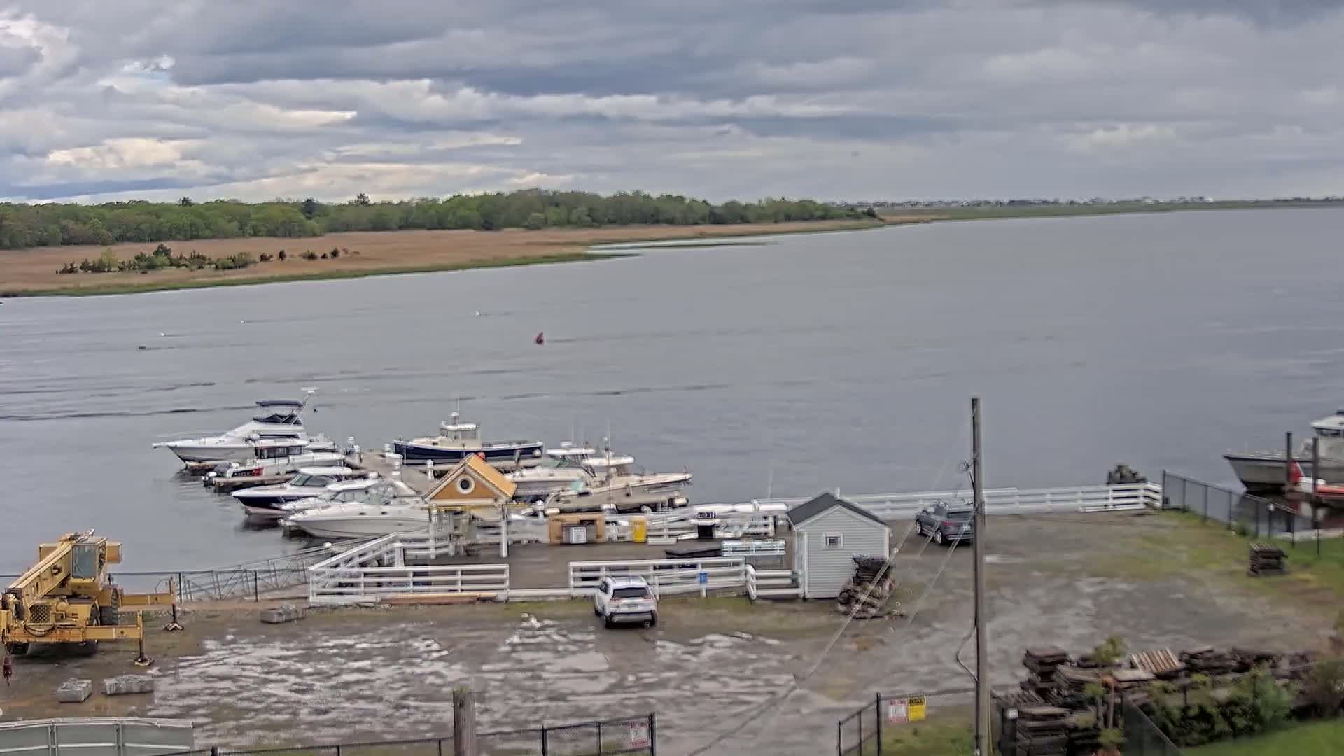 Several boats are docked at a marina on a calm, overcast day next to a small, fenced-in area with a shed and a vehicle.