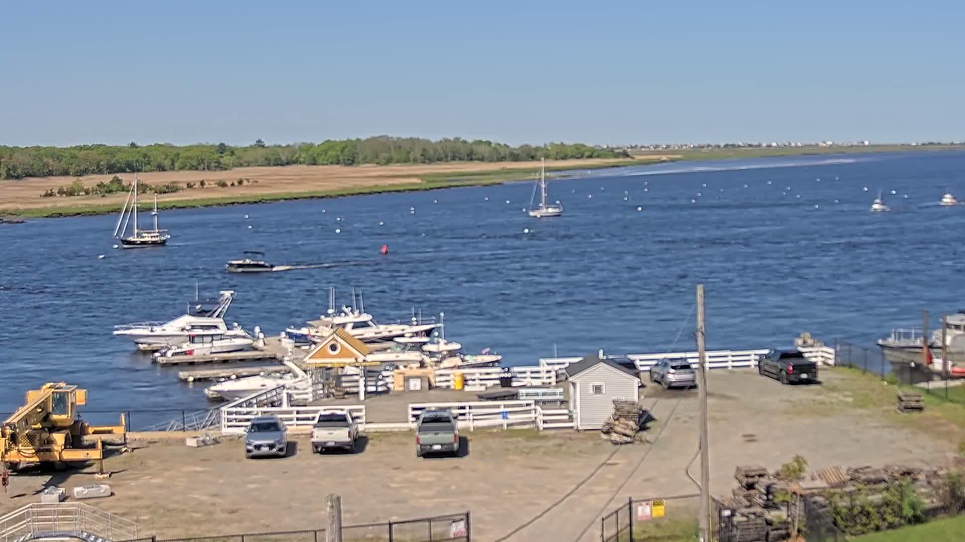 A marina with several boats docked, a few cars parked in a lot, and a large body of water with other boats sailing under a clear blue sky.