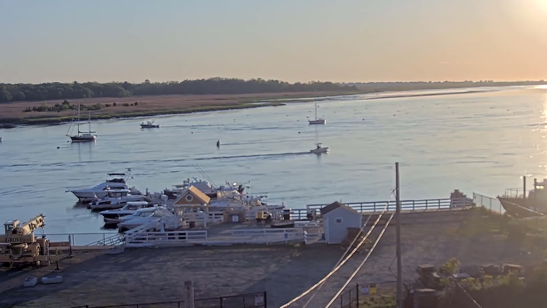 A calm, sunlit body of water with several sailboats and motorboats, some docked at a marina with a small building, and a crane visible on the shore.