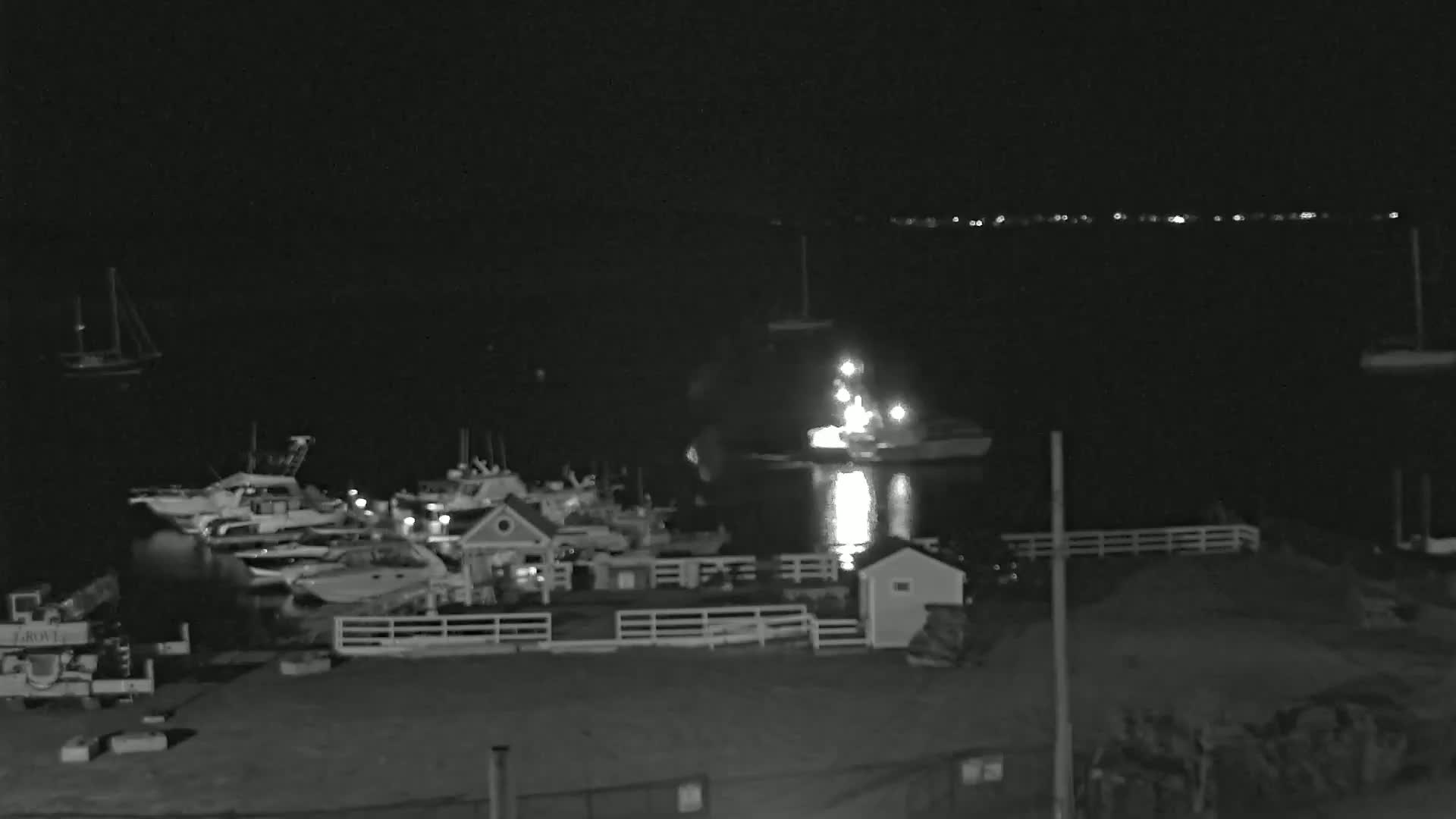 A nighttime, low-light view of a marina with numerous boats docked, a small building, and a brightly lit vessel in the water.