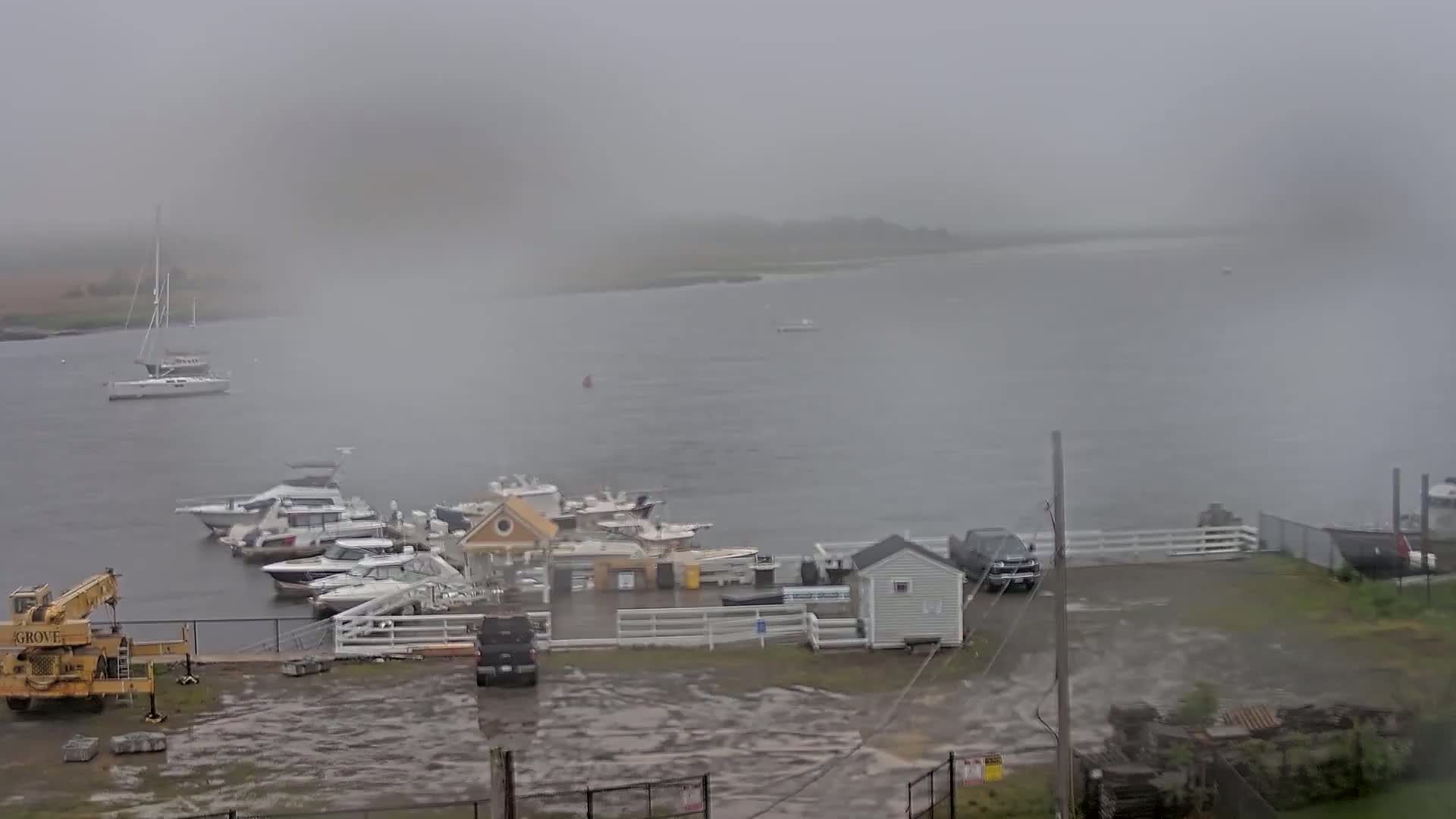 A rainy, hazy day at a marina shows several boats moored, some in the water and others docked, along with a yellow crane and a few vehicles in the parking lot.