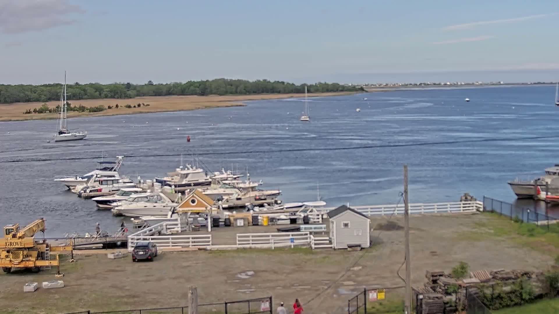 A marina with numerous boats docked, a small building, and a crane is situated on a calm body of water under a clear sky.
