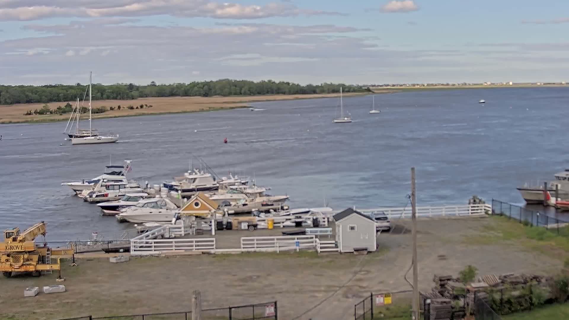 A marina with several boats docked, including sailboats and motorboats, sits on a calm, gray-water river under a partly cloudy sky.