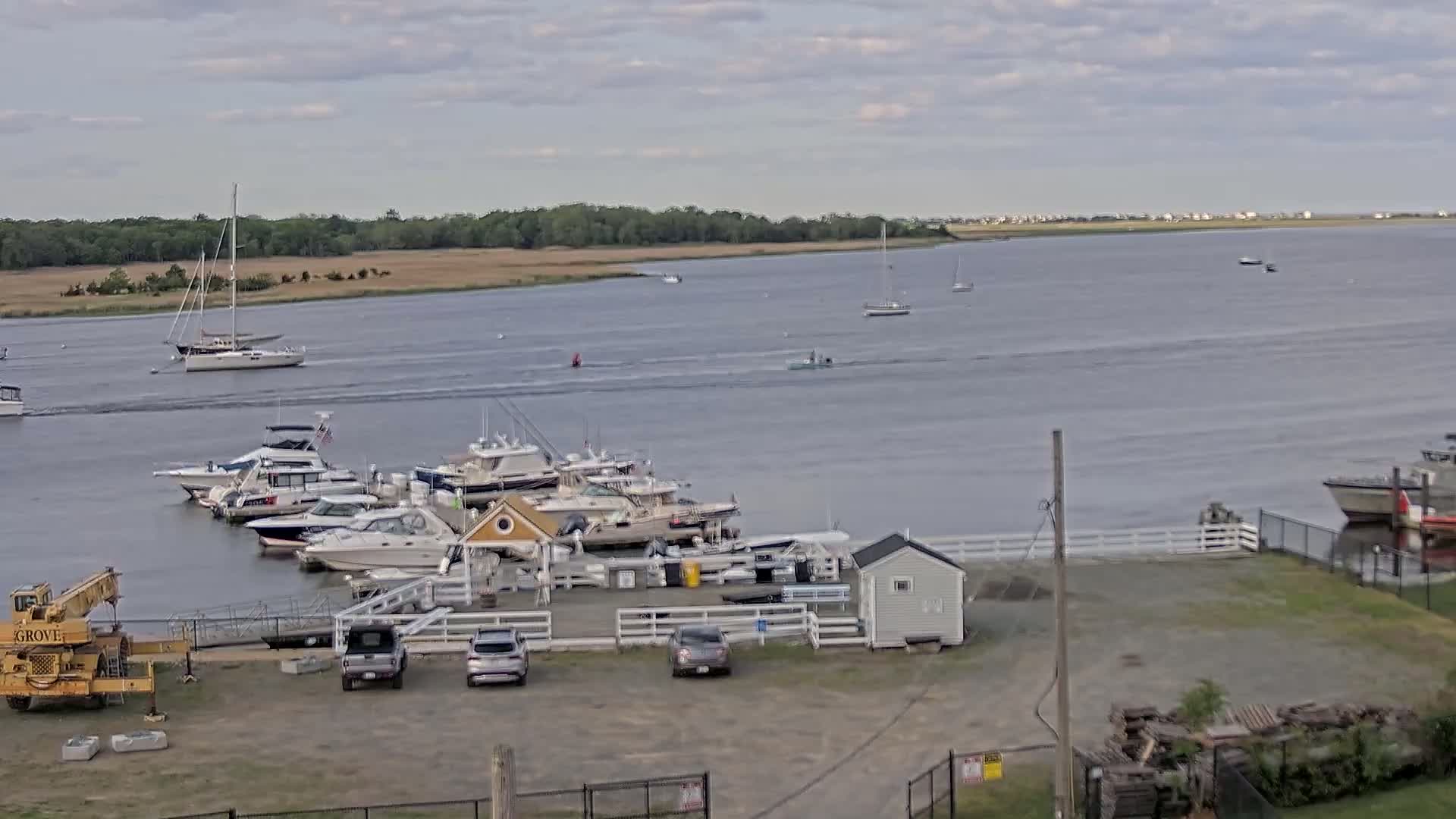 A marina with several boats docked, a few cars in a parking lot, and a large body of water with additional boats and sailboats under a cloudy sky.