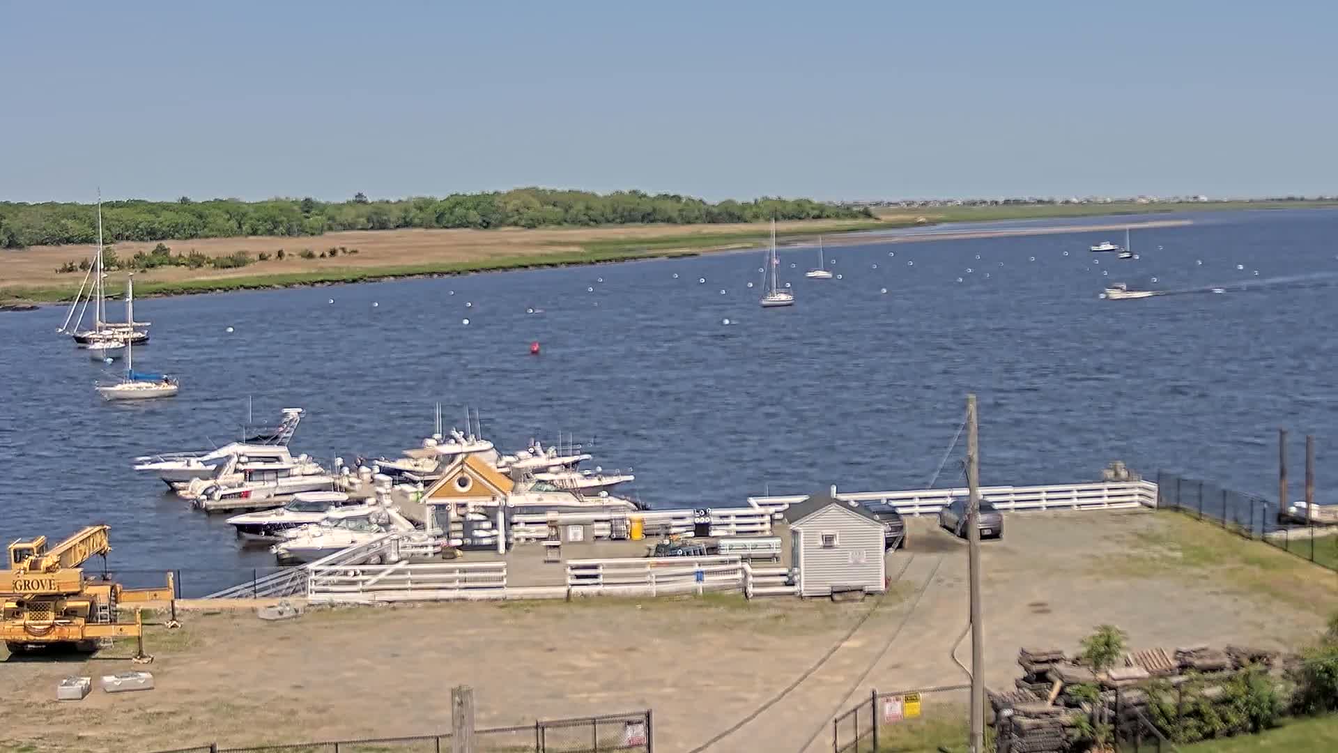 A marina with several boats docked on a calm, sunny day, overlooks a body of water with additional boats and a shoreline of trees and marshland.