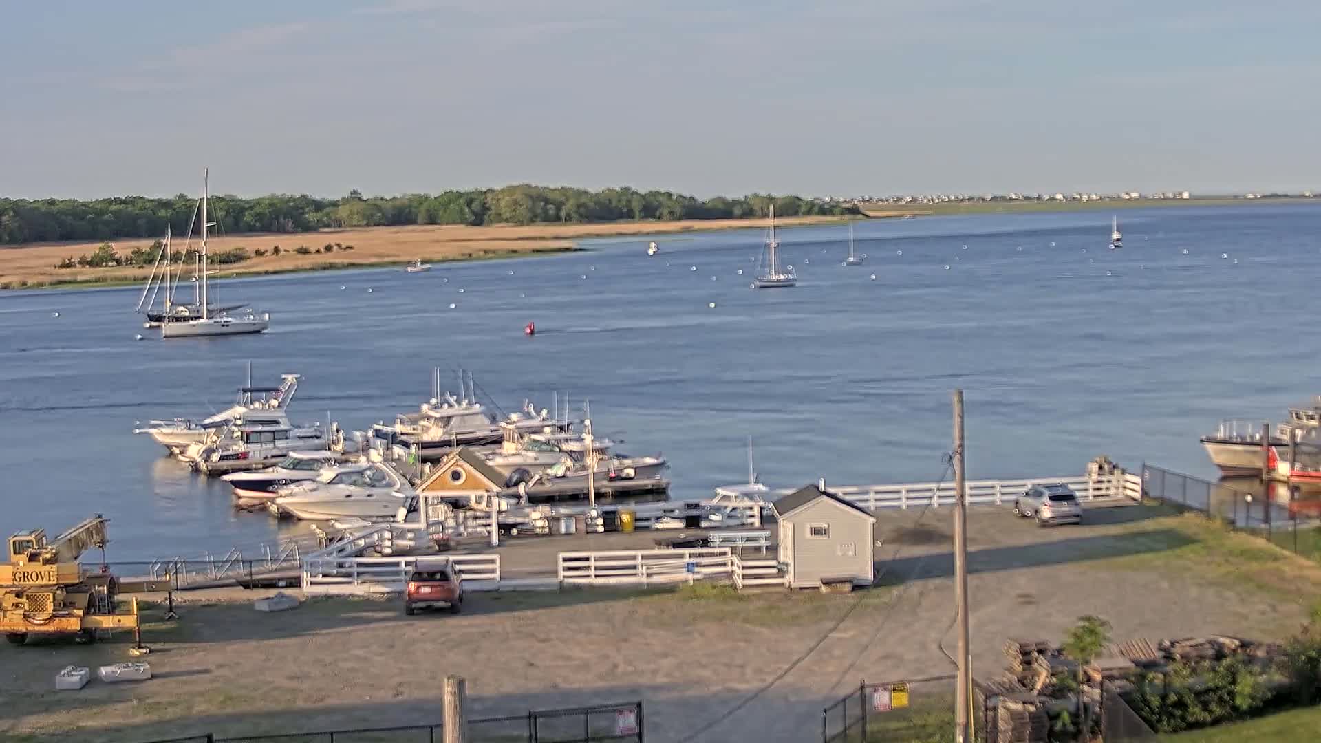 A calm, sunny day shows a marina with several motorboats docked, a few sailboats on the water, and a shoreline with trees in the background.