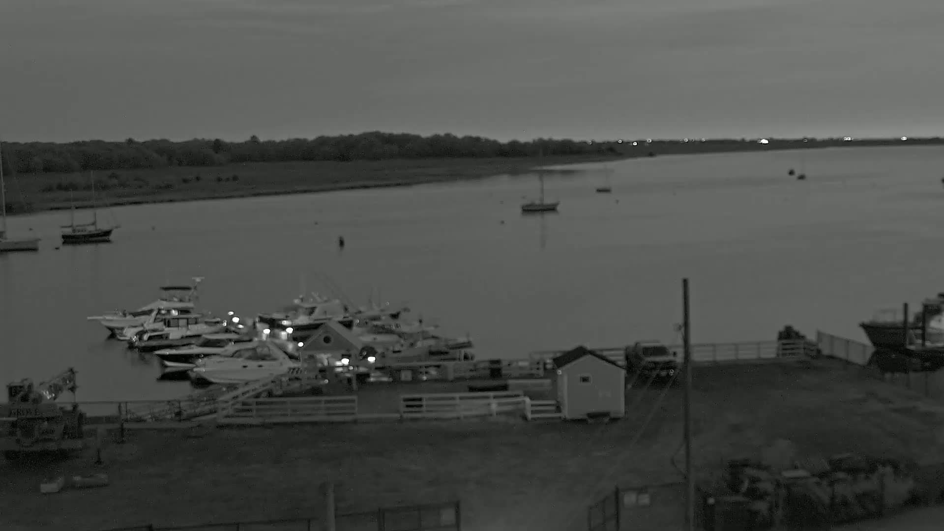 A marina with numerous boats moored at dusk under a cloudy sky, adjacent to a calm body of water with a few more boats visible further out.