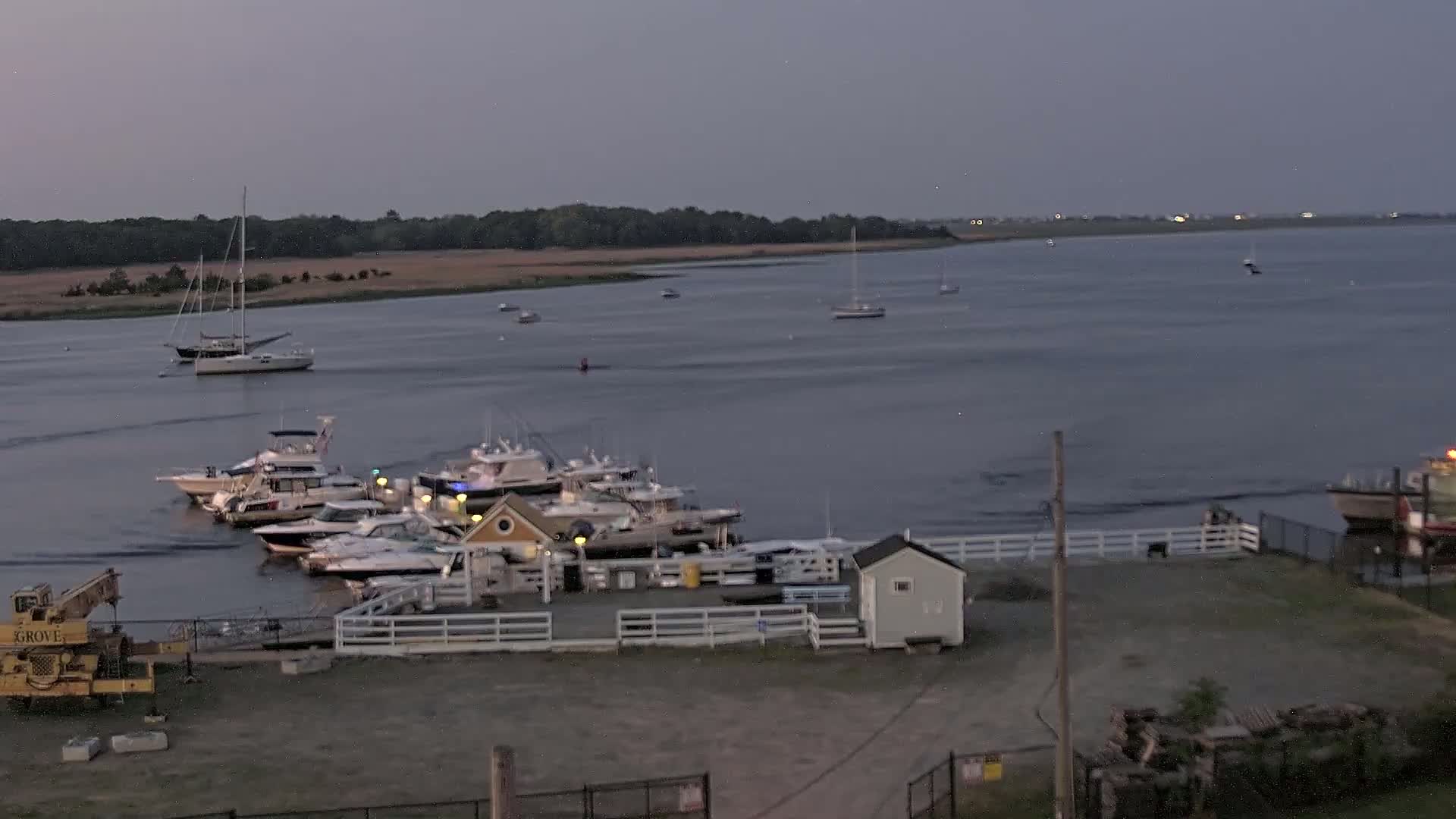 A marina filled with motorboats and sailboats sits on the waterfront at dusk, under a cloudy sky.