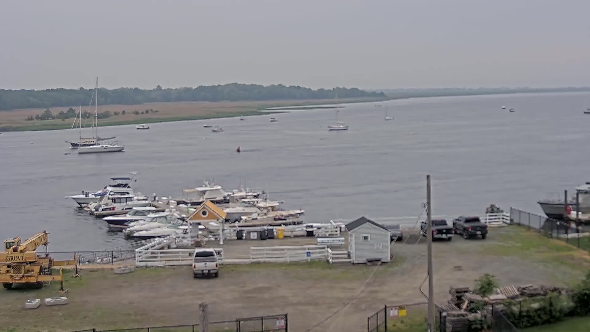 A marina with numerous boats docked, a few cars parked in a lot, and a calm body of water with several more boats in the distance under an overcast sky.