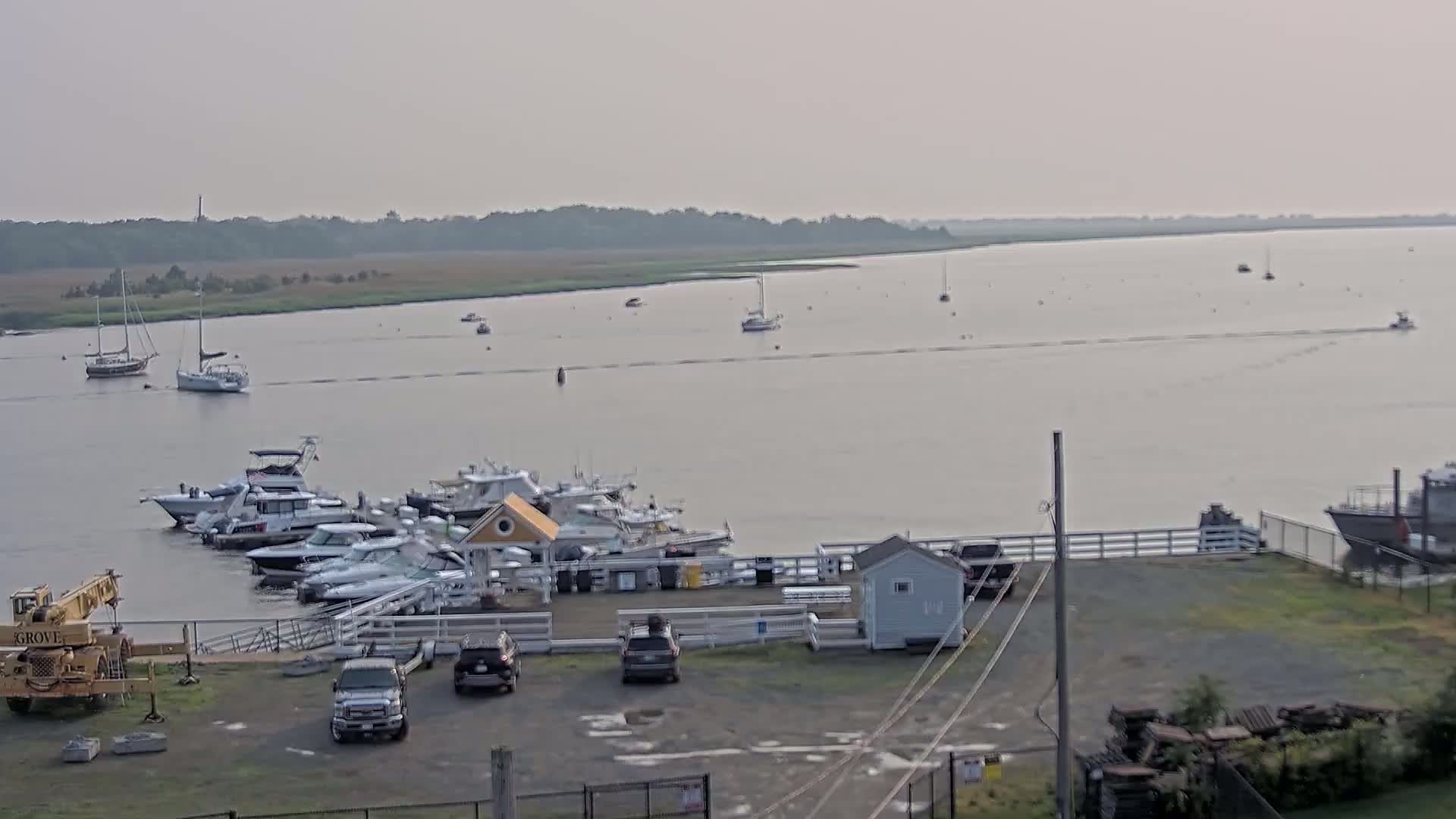 A calm, hazy day reveals a marina filled with boats,  with several more boats and sailboats visible on the wide waterway beyond.