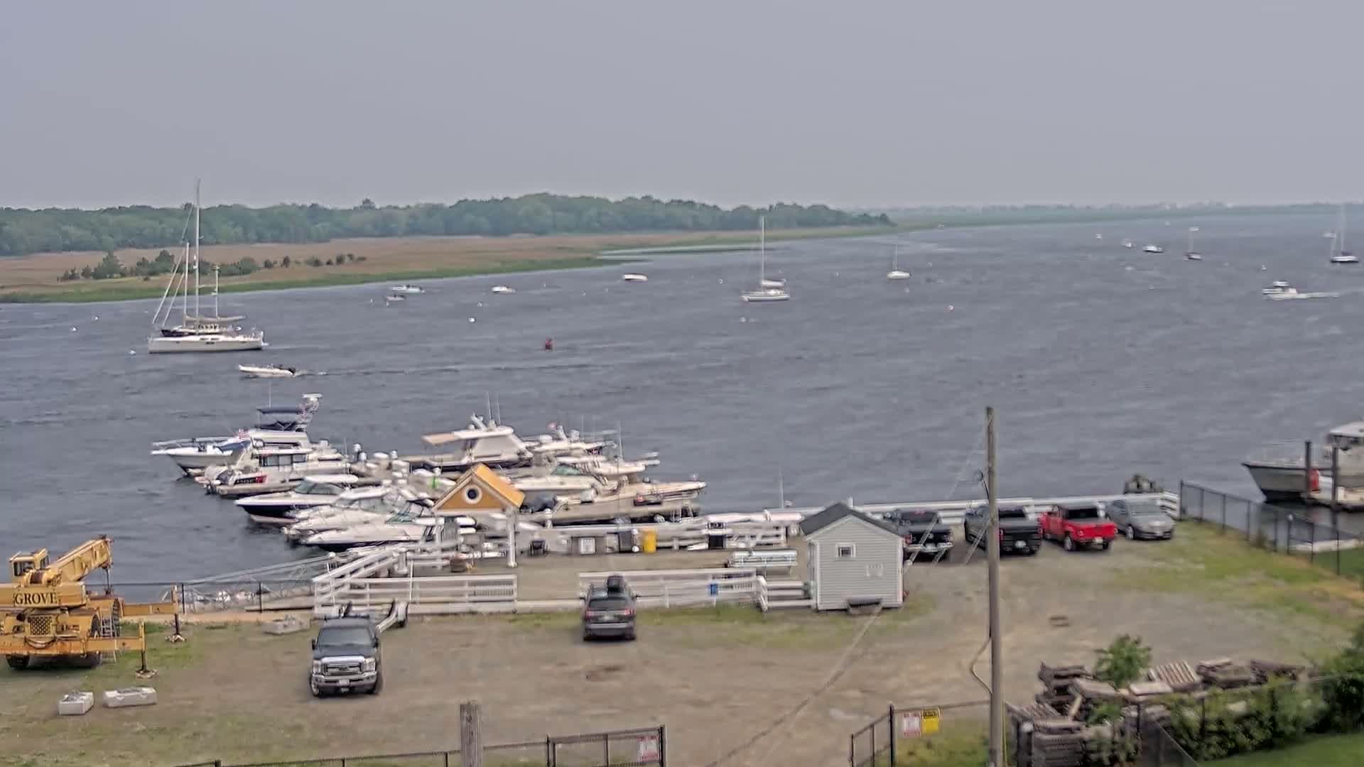 A marina with numerous boats moored, some on the water and others docked, is situated on a shoreline with a slightly choppy body of water under an overcast sky.