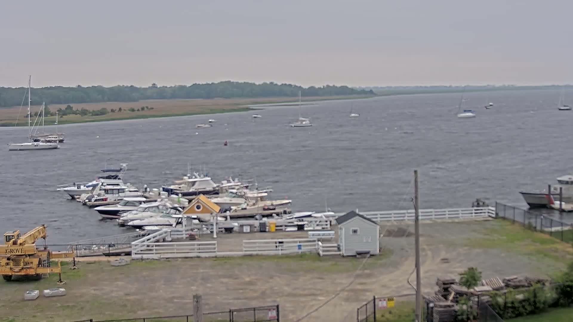 A marina with numerous boats moored, situated on a windy, overcast day next to a body of water with additional boats visible in the distance.