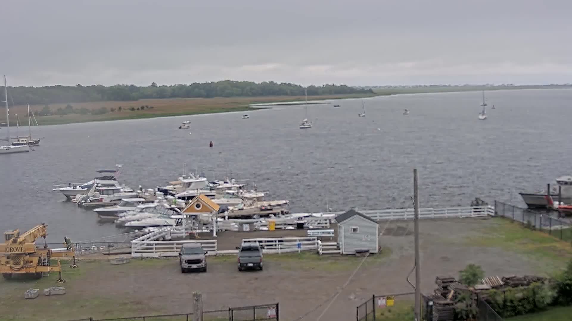 A marina with numerous boats docked under an overcast sky, with several more boats visible on the choppy water in the background.