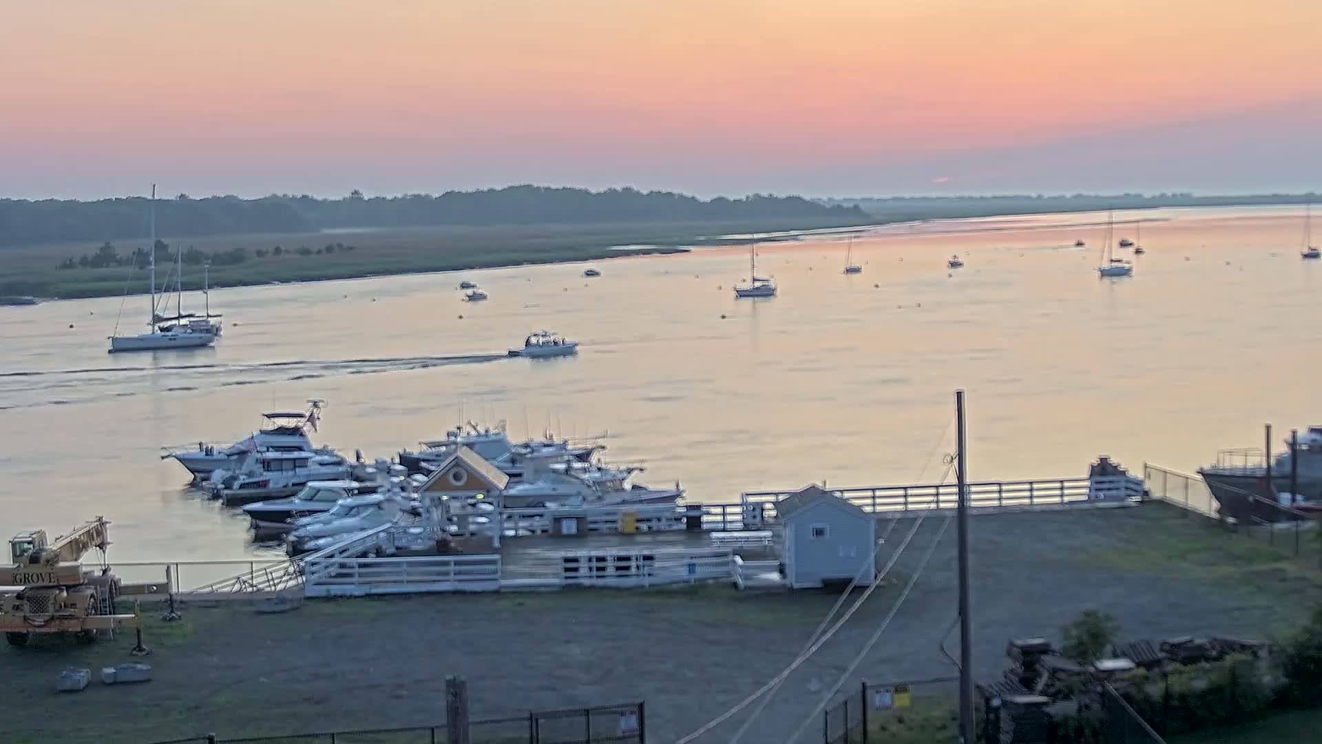 Several boats are docked at a marina during a calm, pinkish sunrise over a wide river.