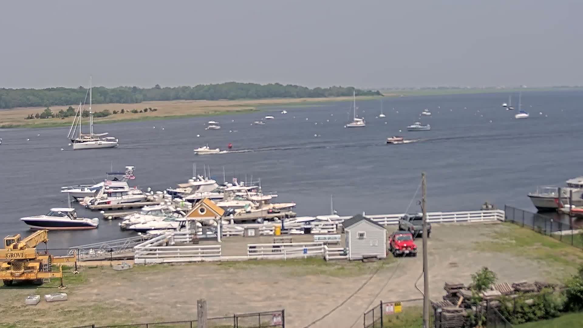A marina filled with numerous boats and yachts is situated on a calm, gray body of water under a hazy sky, with additional boats visible in the distance.