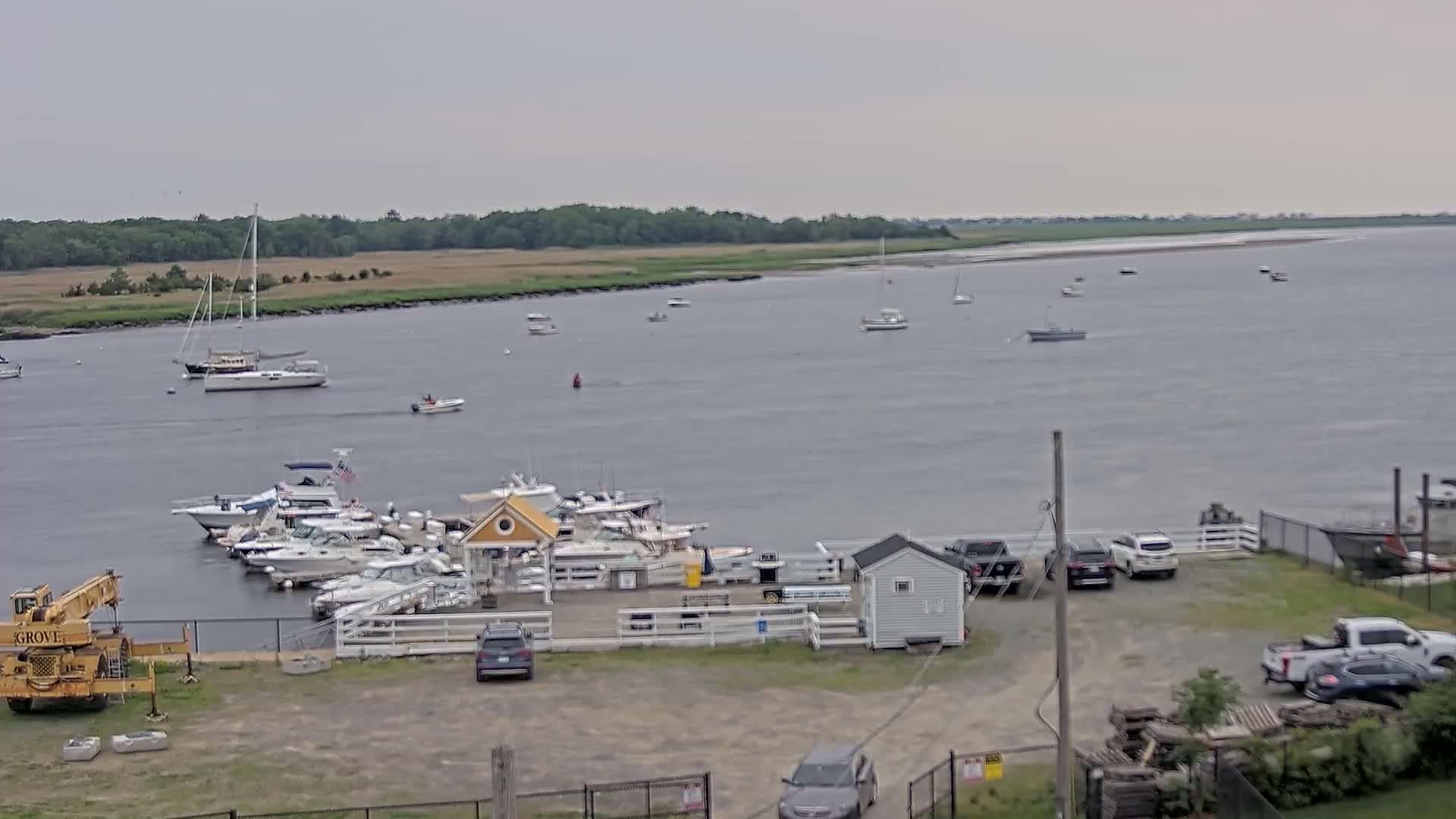 A marina with numerous boats docked,  cars in a parking lot, and a view of a calm body of water under an overcast sky.