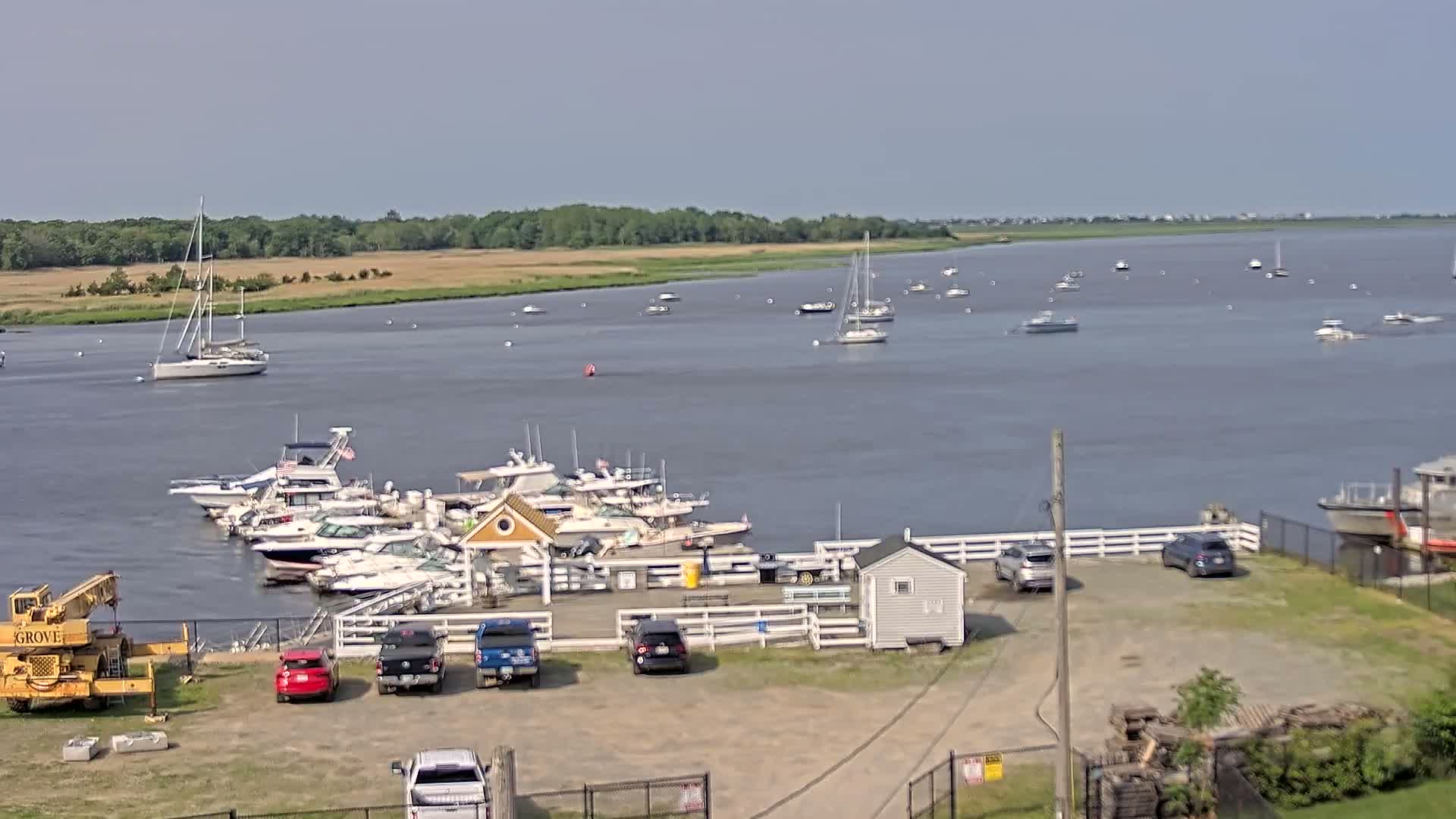 A marina with numerous boats docked, several cars parked nearby, and a calm body of water with additional boats scattered across its surface under a mostly clear sky.