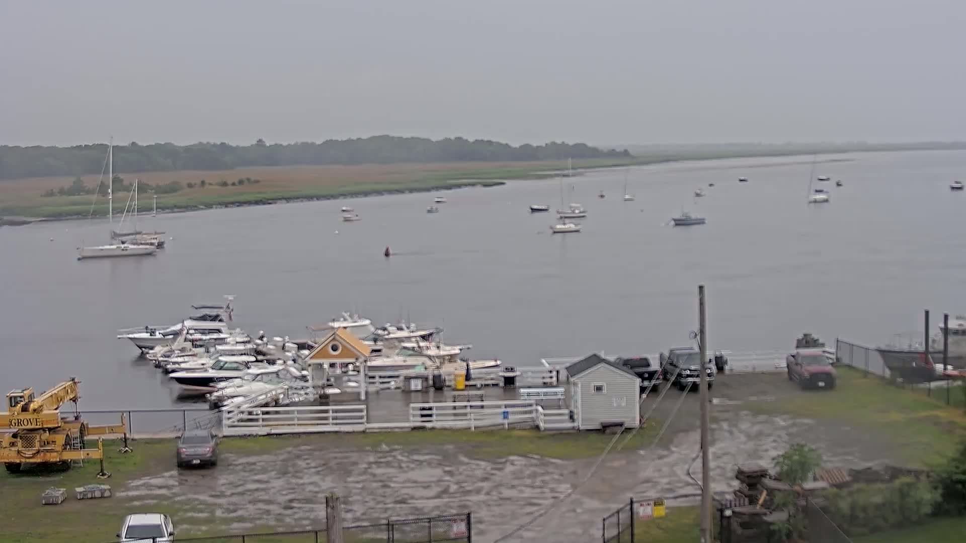 A marina with numerous boats docked, some additional boats on the water, and a few vehicles parked in a lot under an overcast sky.