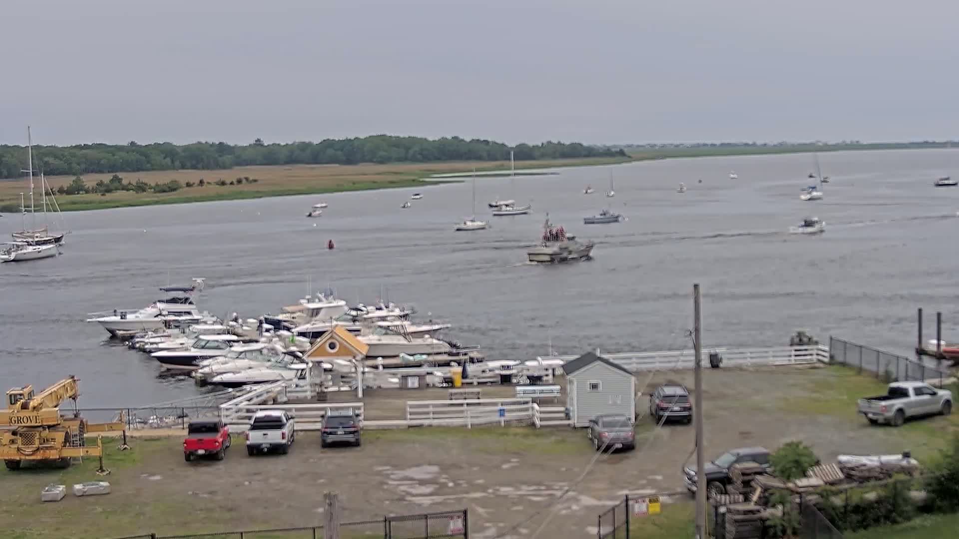 A marina with numerous boats docked, a few cars parked in a lot, and a body of water with several more boats under an overcast sky.