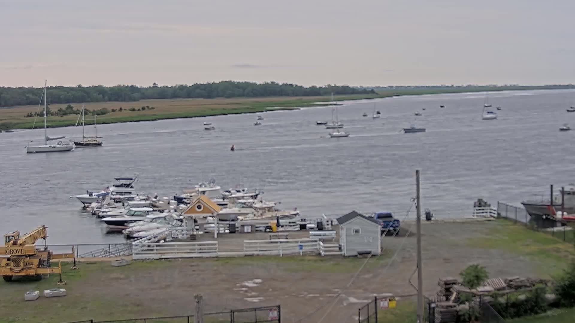 A marina with numerous boats docked, a few cars parked in a lot, and a body of water with several more boats under an overcast sky.