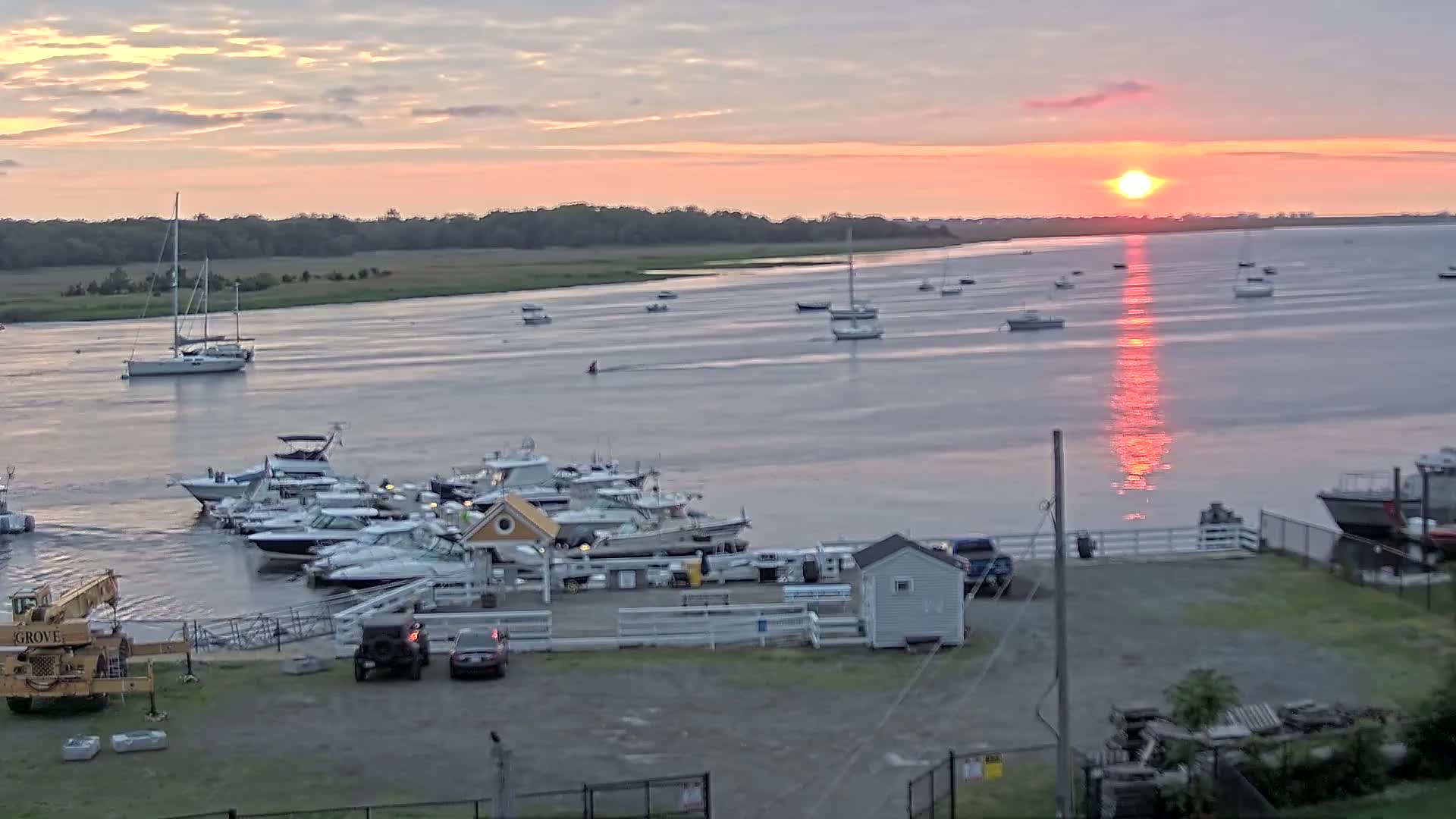 A calm body of water with numerous boats at a marina is seen at sunset, with the sun reflecting brightly on the water's surface.