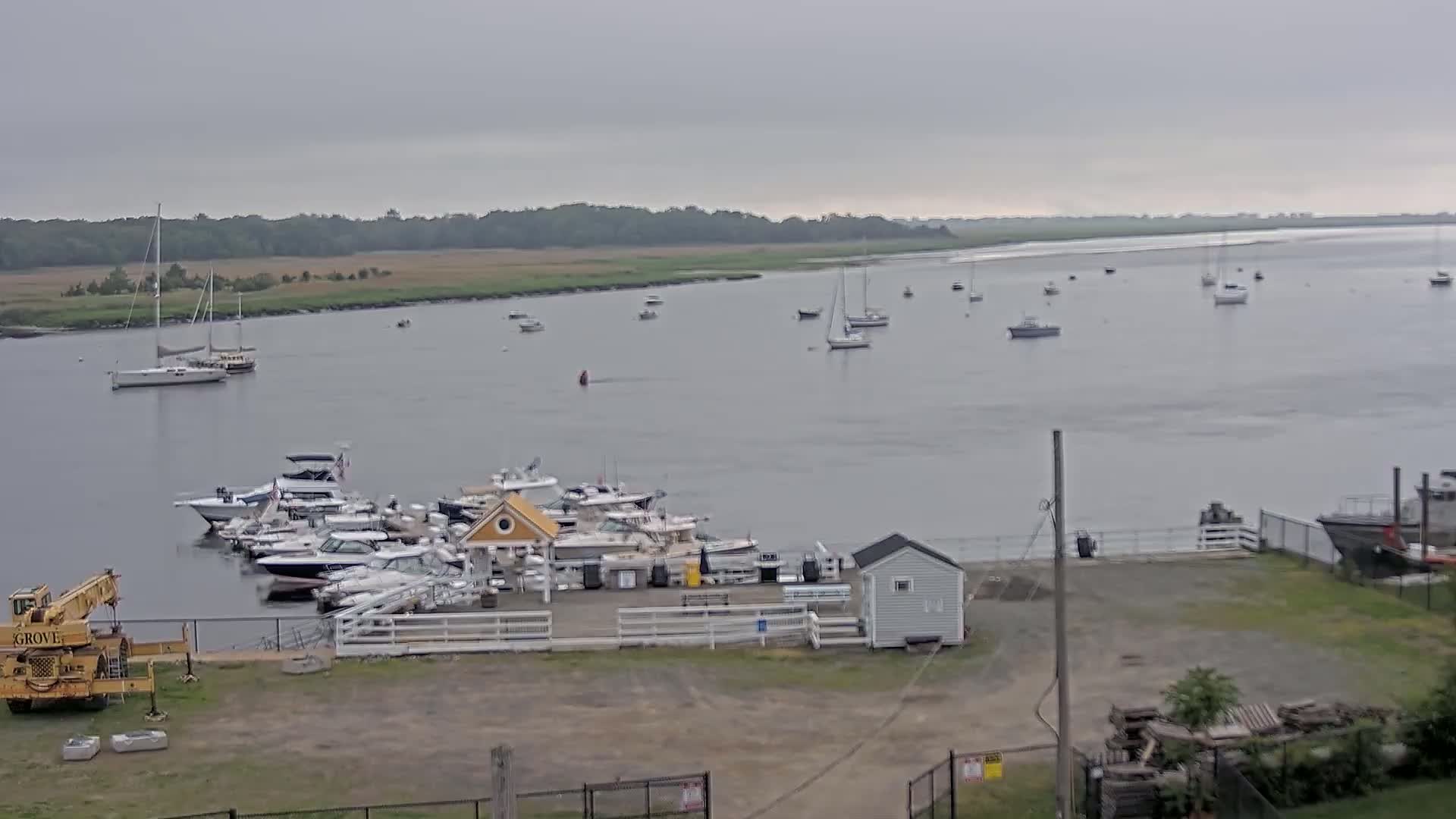 A marina with numerous boats docked, situated on a calm body of water under an overcast sky, with additional boats visible further out in the bay.