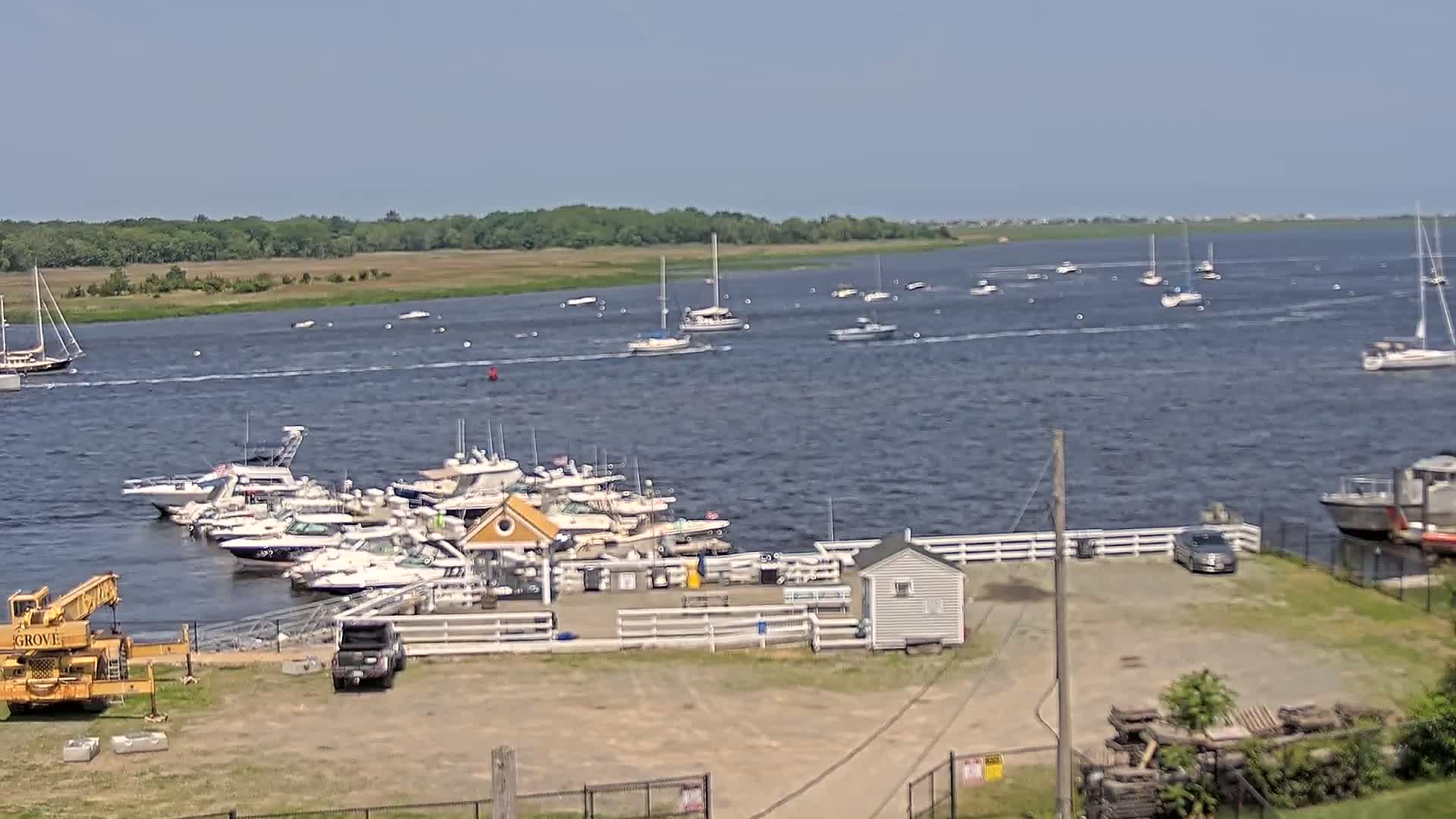 A marina with numerous boats docked, a yellow crane, and several small buildings is situated on a shoreline overlooking a calm body of water with scattered sailboats and motorboats under a clear sky.