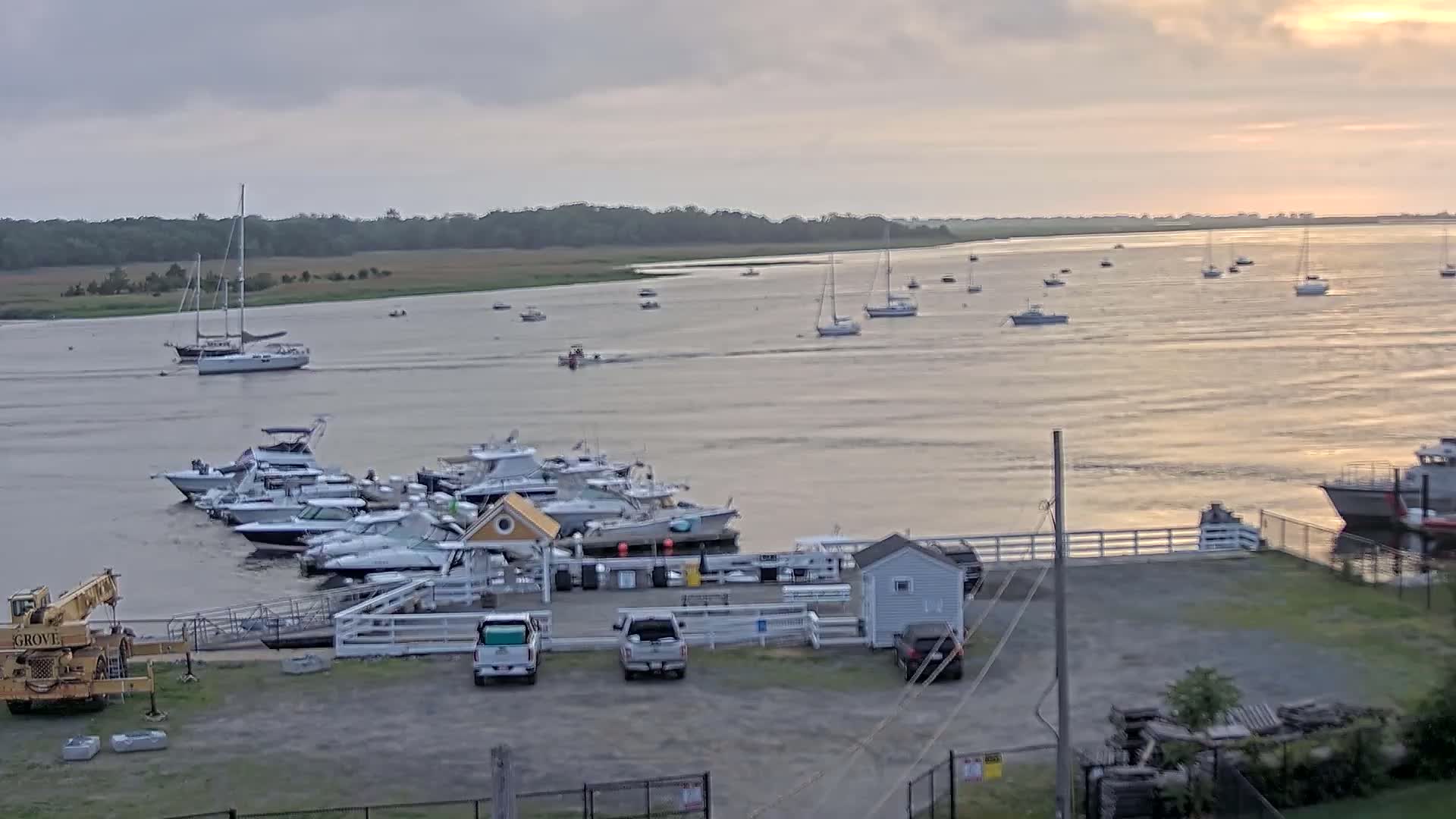 Numerous boats are docked at a marina on a calm, overcast day, with additional boats visible on the water beyond.