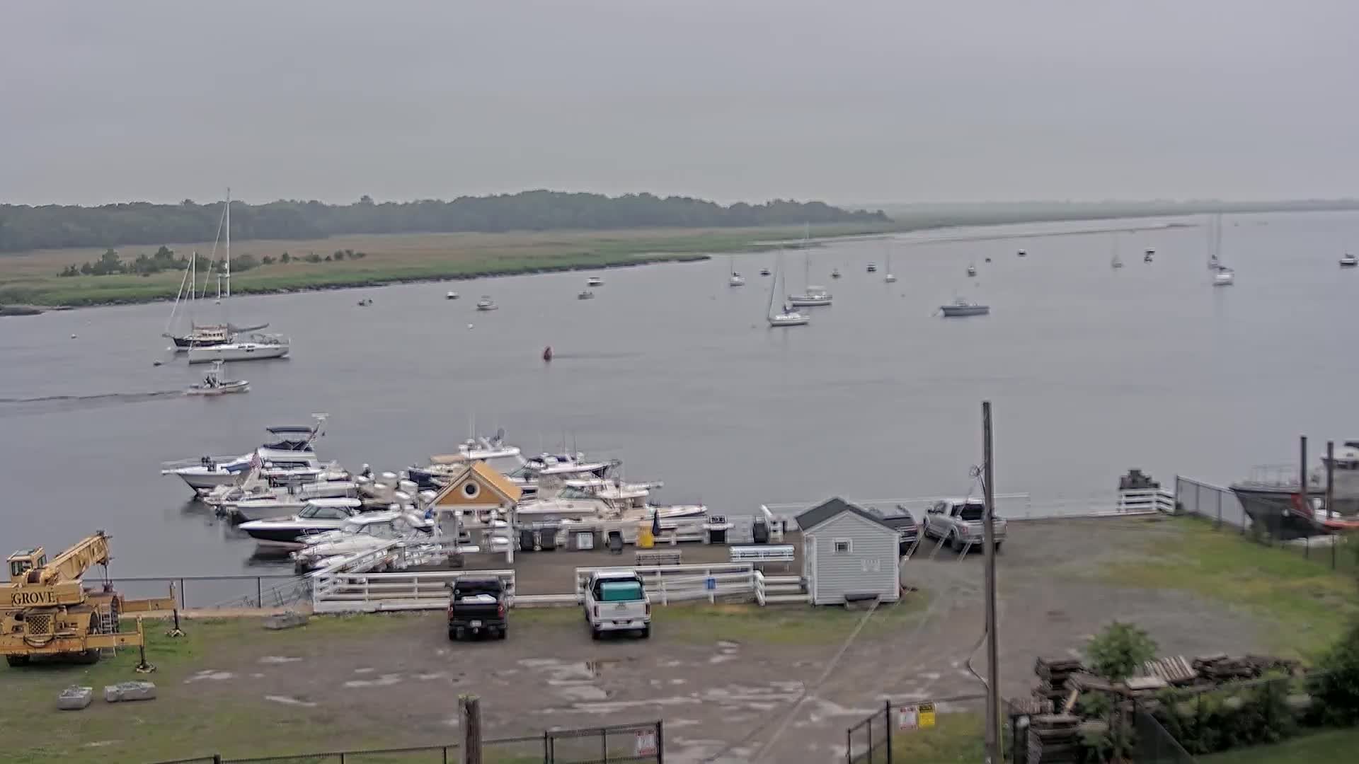 A marina with numerous boats docked, along with several vehicles in a parking lot, is shown on an overcast day, overlooking a calm body of water with additional boats and a shoreline of greenery in the background.